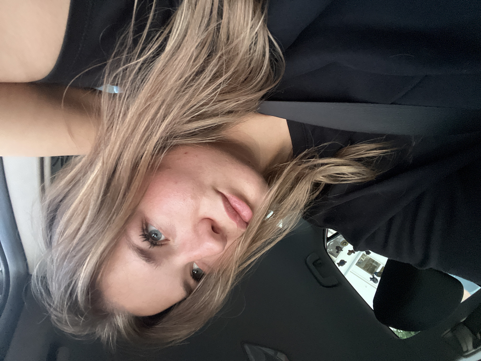 The image depicts a young woman with light brown hair and fair skin in what appears to be the interior of a car. She is wearing a black shirt and a seatbelt is visible. The background shows parts of the car interior, including the dashboard and a glimpse of the outside through the window. It is not possible to determine if the location is in Budva, Montenegro from the given image alone.