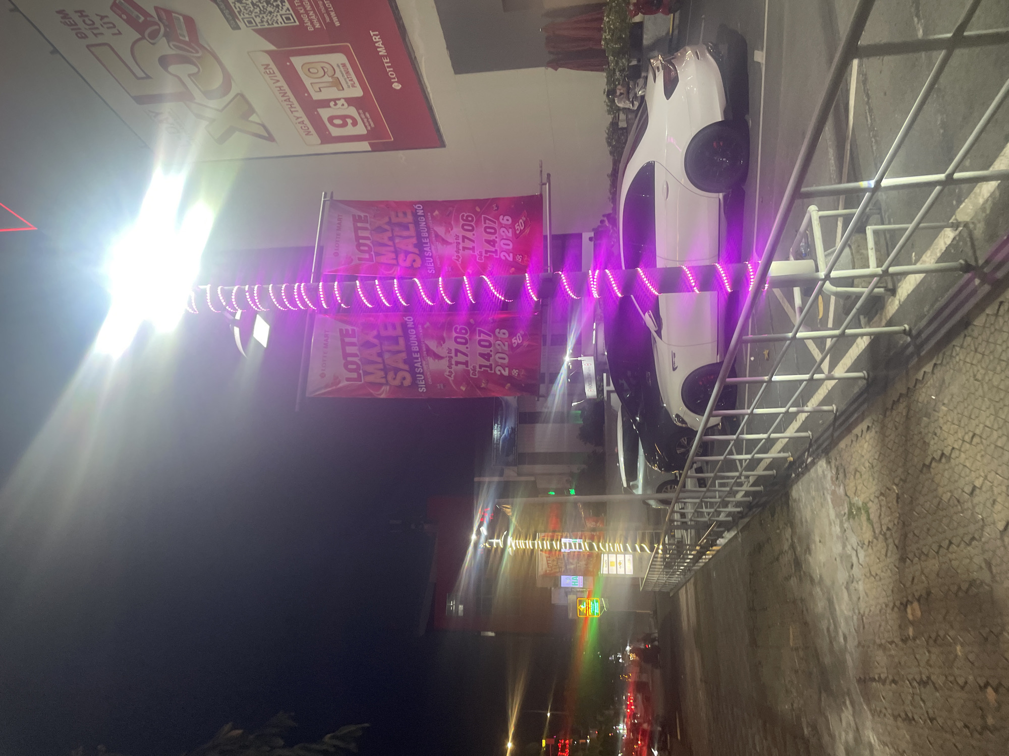 The image captures a nighttime scene outside a Lotte Mart in Thuận An, Vietnam. A lamppost wrapped in purple LED lights stands prominently in the foreground, illuminating two large banners advertising a "LOTTE MAX SALE." The banners indicate the sale is effective from 17.06 to 14.07, 2026, with "SIÊU SALE BUNG NỔ" meaning "Explosive Super Sale." To the right of the banners, a large red sign displays "X5" and "NGÀY THÀNH VIÊN," suggesting a membership bonus.
In the parking lot, a white sedan and a dark-colored sedan are parked. A few people are visible, including one individual sitting on a scooter in the background and another person standing near plants to the right. Traffic is visible on a street to the left, with blurred red and white lights indicating moving vehicles. The ground is paved with cobblestones and concrete. The overall atmosphere suggests a commercial area at night, likely a shopping mall or department store with promotional activities.
