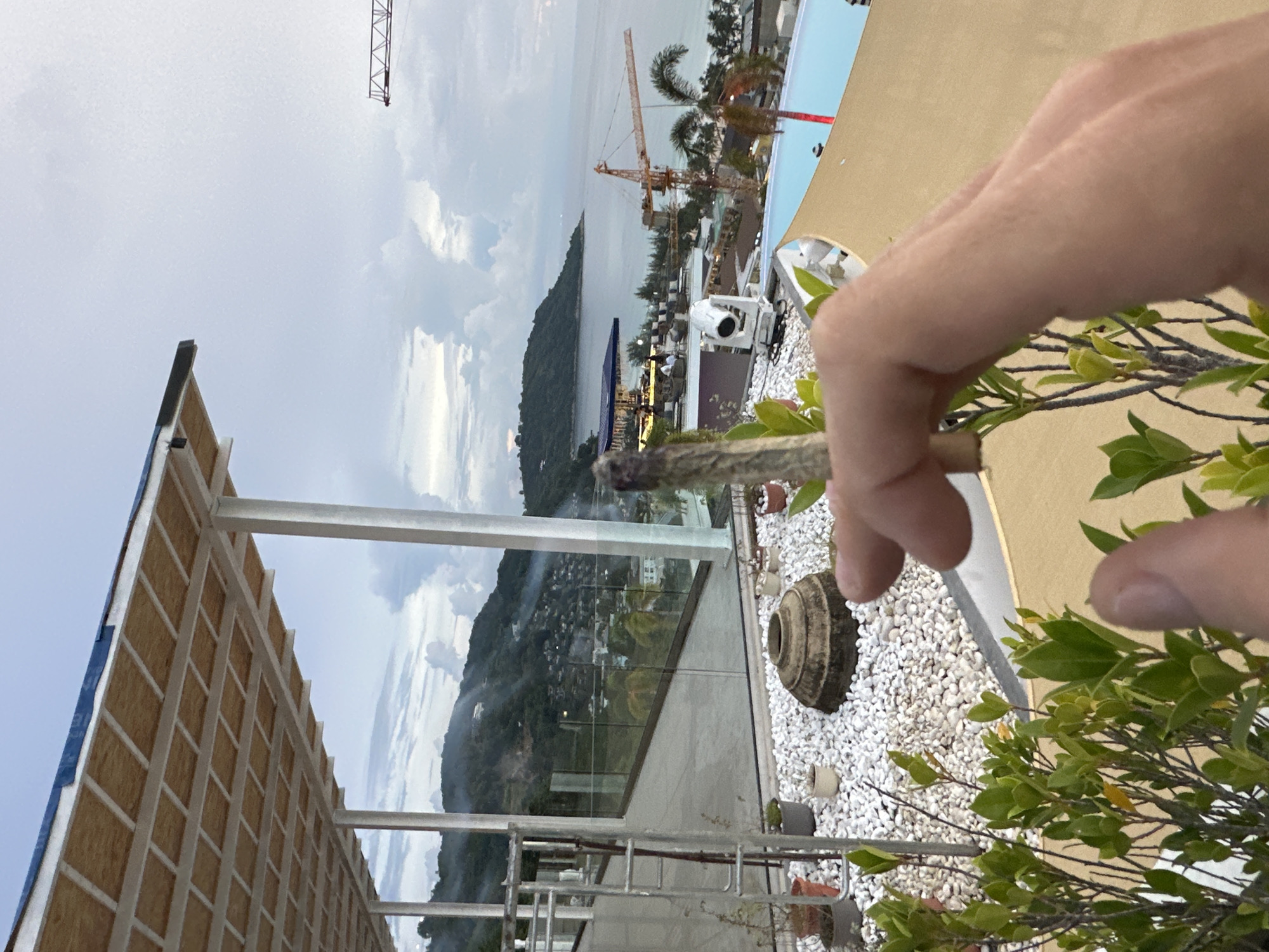 The image depicts a hand holding a cannabis joint in the foreground, with a hazy view of Karon, Thailand, in the background. The scene appears to be from a balcony or rooftop, overlooking a coastal area. In the distance, hills covered with vegetation and scattered buildings are visible. The sea stretches out towards the horizon, under a cloudy sky. Construction cranes are present, indicating ongoing development in the area. A swimming pool and outdoor seating area are also visible, suggesting a resort or hotel setting. The overall atmosphere is casual, with the focus on the hand and the joint.
