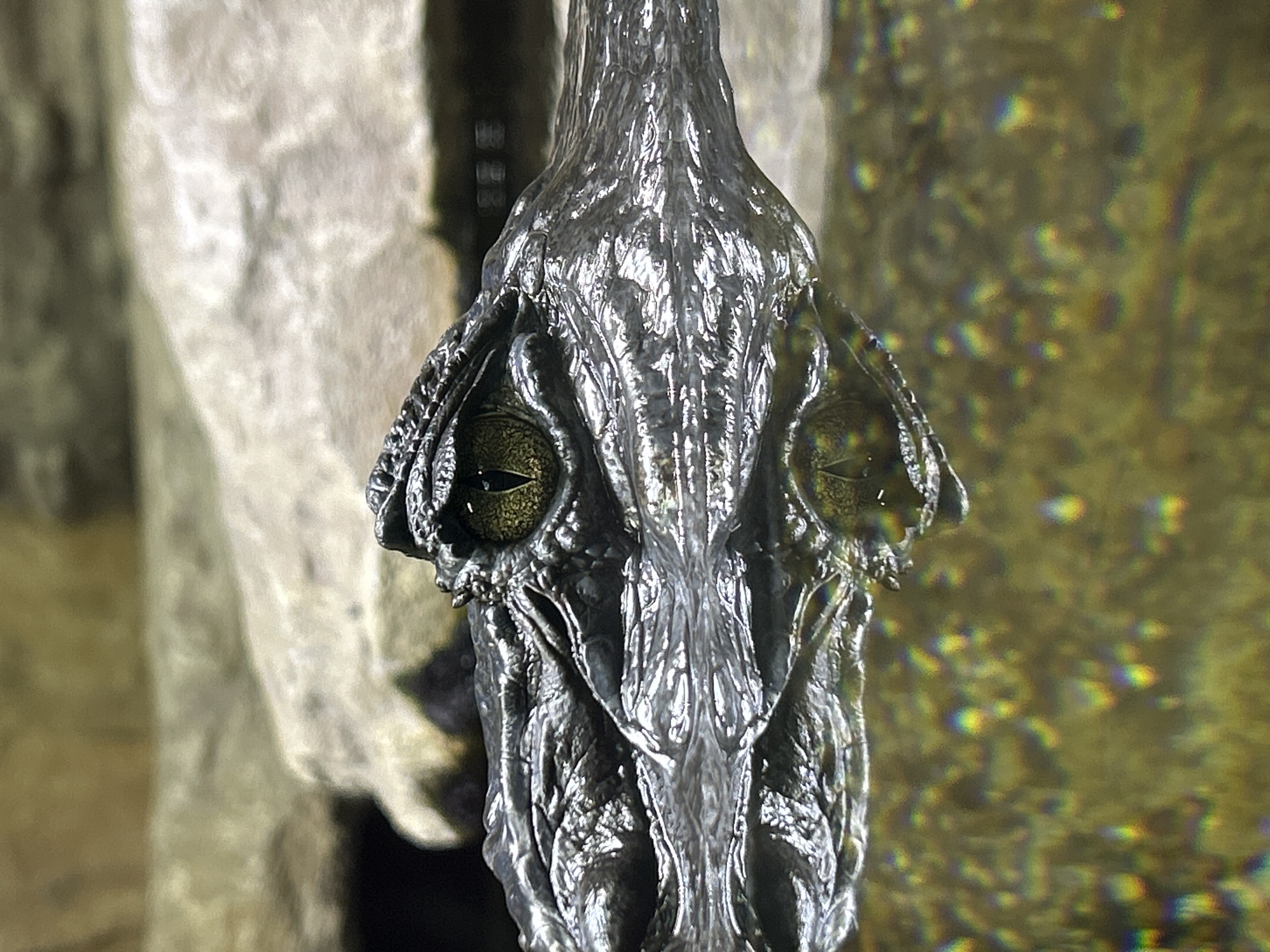 A close-up, eye-level shot captures an alligator's eye and snout partially submerged in murky water. The alligator's rough, scaly skin is detailed and reflects the light. Its eye, a golden-yellow iris with a dark vertical pupil, is partially closed, giving it a watchful appearance. The water's surface is disturbed, with ripples distorting the reflection of the alligator. Sunlight refracts through the water, creating a spectrum of rainbow-like colors scattered across the bottom of the frame. The background consists of indistinct, rocky formations, suggesting a natural or simulated habitat. There is no indication of any specific location in Yekaterinburg, Russia.