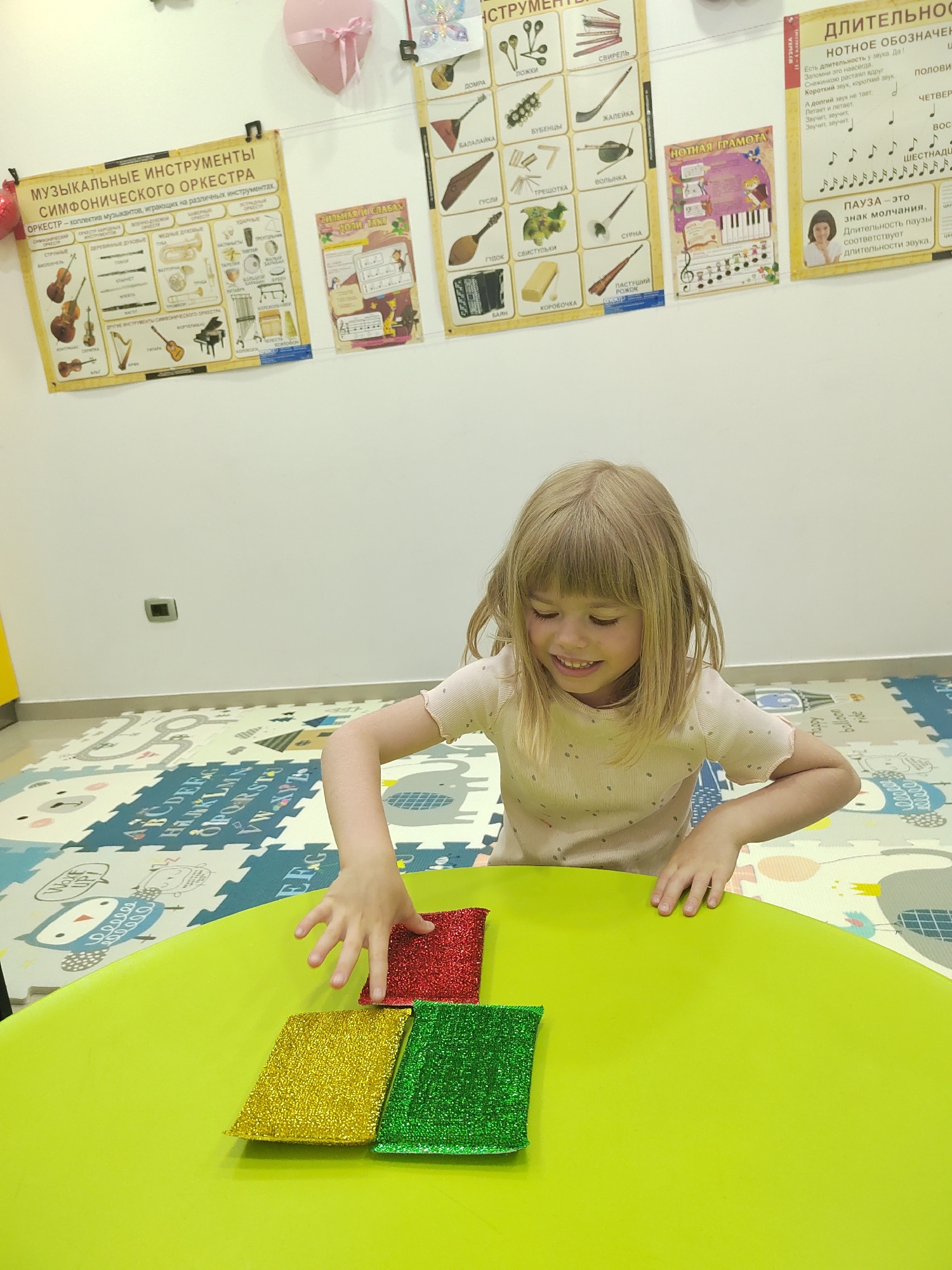 A young girl with blonde hair and a light-colored, short-sleeved top is captured smiling as she reaches for colorful, sparkly objects on a bright green table. She is looking down, engaging with three square-shaped, glittery cushions or beanbags in red, yellow, and green.
The scene takes place indoors, likely in a playroom or educational setting, possibly a music classroom, in Podgorica, Montenegro. The floor is covered with interlocking foam mats featuring alphabet letters (e.g., A, B, C, D, E, F, G, H, I, J, K, L, M, N, O, P, Q, R, S, T, U, V, W, X, Y, Z are visible) and cartoon animal designs, including an owl and an elephant. The walls are white and adorned with several educational posters related to music.
Visible text on the posters includes:
* "МУЗЫКАЛЬНЫЕ ИНСТРУМЕНТЫ СИМФОНИЧЕСКОГО ОРКЕСТРА" (Musical Instruments of the Symphony Orchestra), which defines an orchestra and categorizes instruments into strings, woodwinds, brass, percussion, and others, showing illustrations of instruments like violins, harp, piano, trumpet, and various drums.
* Smaller posters display traditional and folk instruments such as "ДОМРА", "БАЛАЛАЙКА", "ГУСЛИ", "ГУДОК", "БАЯН", "ЛОЖКИ" (spoons), "СВИРЕЛЬ" (pipe), "БУБЕНЦЫ" (bells), "ЖАЛЕЙКА" (hornpipe), "ТРЕЩОТКА" (ratchet), "ВОЛЫНКА" (bagpipe), "СВИСТУЛЬКИ" (whistles), "КОРОБОЧКА" (wood block), and "ПАСТУШИЙ РОЖОК" (shepherd's horn).
* Another poster titled "НОТНАЯ ГРАМОТА" (Musical Notation) shows musical staff with notes and piano keys.
* A poster about "ДЛИТЕЛЬНОСТЬ НОТНОЕ ОБОЗНАЧЕНИЕ" (Duration Musical Notation) explains various note durations (whole, half, quarter, eighth, sixteenth) and states "ПАУЗА - это знак молчания" (REST - is a sign of silence).
A pink heart-shaped decoration hangs on the wall near the top left corner. The lighting suggests an indoor setting with no specific time of day discernible. The girl appears happy and engaged in her activity.