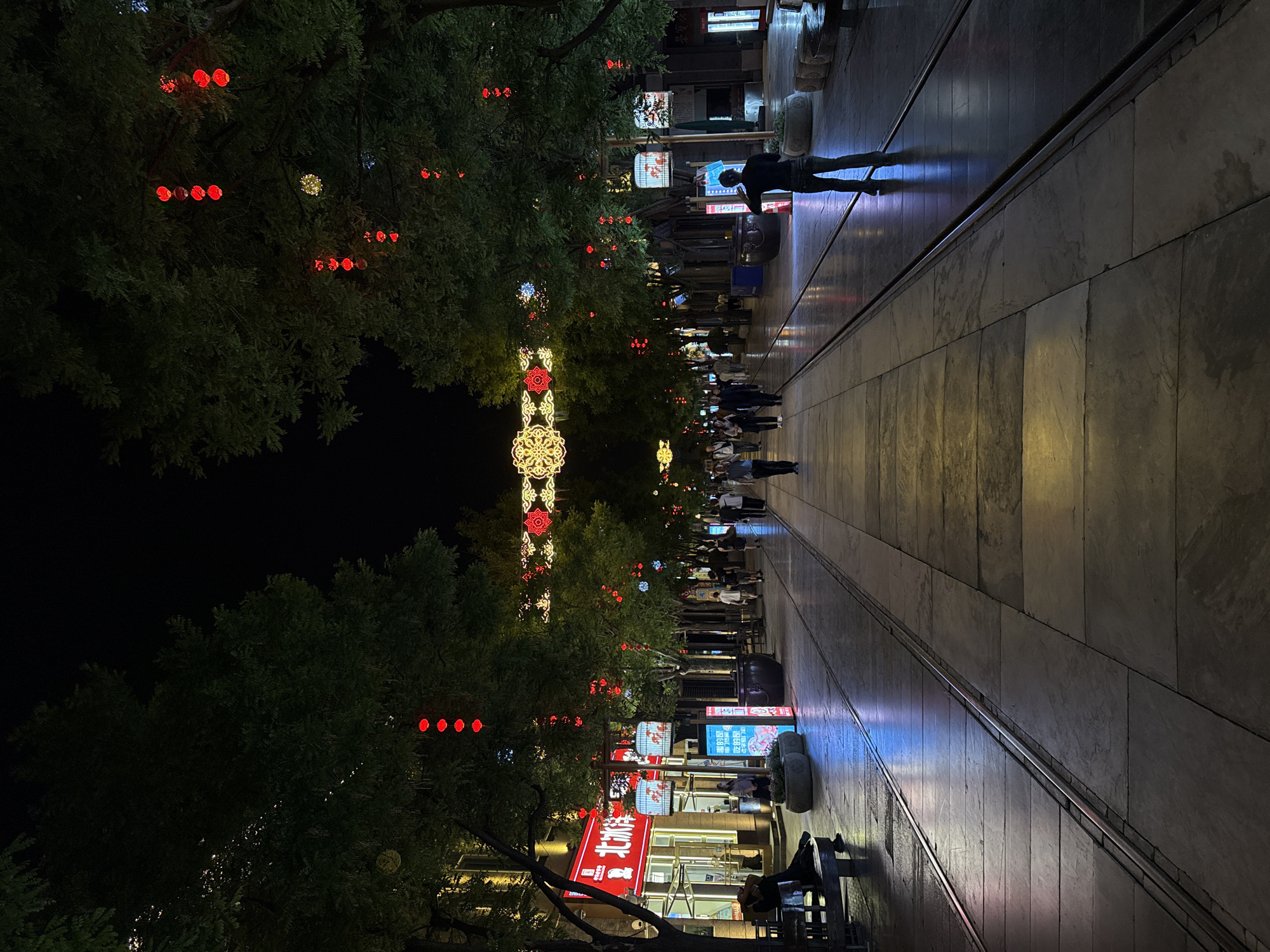 The image shows a brightly lit pedestrian street at night in the Dongcheng District of Beijing, China. The street is lined with trees decorated with red and white lights and ornate illuminated structures. People are walking along the paved pathway. The street has a modern aesthetic, with tall buildings visible on either side, lit by various signs and displays. Some people are sitting on benches. The ground is made of large, light-colored stone slabs.