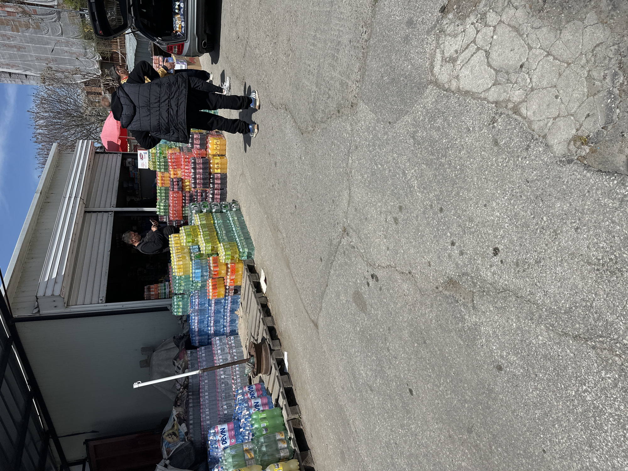 The image captures an outdoor commercial scene in bright daylight, likely a wholesale beverage distributor or warehouse. Numerous plastic bottles of various colored drinks, including water, juices, and soft drinks, are stacked high on wooden pallets outside a building. Brands like "DEVIN" are clearly visible on many of the water bottles, and a white sign with "Coca-Cola" and "Kambanari" in red text is also present.
In the foreground, a person wearing a black jacket and pants, with blue and white sneakers, stands facing away from the camera, next to the open trunk of a silver car. Another individual is partially visible behind them, appearing to interact with the car's trunk, suggesting loading or unloading goods. Inside the building, which has a white facade and a partially rolled-up metal shutter, a person with dark hair is seen looking out, while another figure is visible further inside.
The ground is paved with asphalt, showing cracks and worn patches. In the background, a bare-branched tree stands, and further back, a building covered in scaffolding indicates ongoing construction or renovation. The clear shadows suggest it is a sunny day. The large quantities of bottled beverages and the functional nature of the building point to a commercial activity, possibly distribution or retail of drinks in bulk.