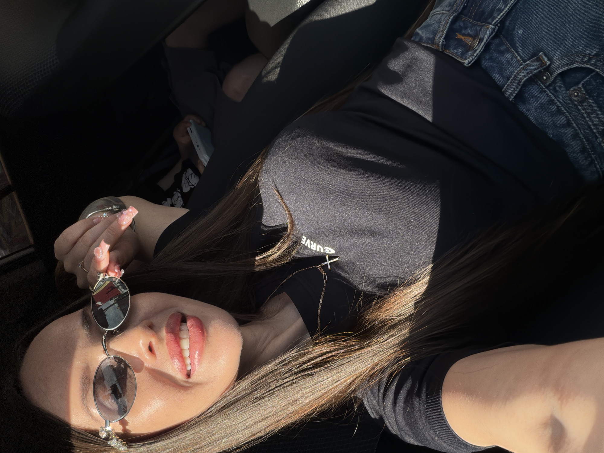 A young woman is depicted reclining horizontally within what appears to be a car interior, under strong, direct sunlight on a bright day in Izhevsk, Russia. Her long, dark brown hair cascades around her, illuminated by the sun.
She is wearing a black t-shirt, on which the partial word "CURVE" is visible in white text, and blue denim jeans. A delicate gold-colored necklace with a small cross pendant adorns her neck. Her left hand, featuring manicured nails with a light, shimmery pink polish, holds a pair of fashionable round-lensed sunglasses with reflective surfaces. Her face, partly in shadow and partly in bright light, shows a relaxed expression with her lips slightly parted in a gentle smile, revealing her teeth and enhanced with glossy lip makeup.
The background indicates an enclosed space, likely a car, with dark interior fabrics and seats visible. In the upper left area, another person's hand or arm is visible, holding a light-colored smartphone that displays a black and white image, possibly featuring Mickey Mouse. The strong sunlight casts sharp shadows across the scene, emphasizing the bright, clear weather conditions. The overall atmosphere conveys a sense of ease and casual travel.