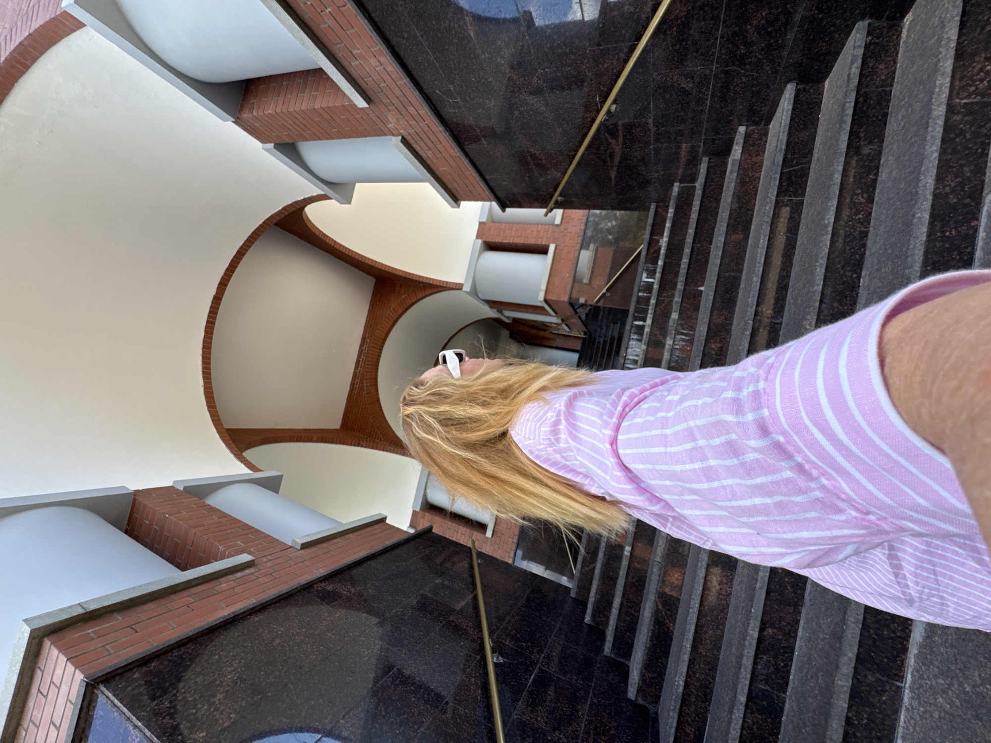 A blonde woman with long, wavy hair is seen from a low angle, walking up a marble staircase. She is wearing a pink and white striped shirt and white-framed sunglasses. The staircase is flanked by dark marble walls and a brick facade. Above, an arched canopy with white trim and brick accents creates a covered walkway. The scene appears to be an outdoor public space, possibly an entrance to a building or park in Saint Petersburg, Russia, characterized by its classical architecture and marble elements.