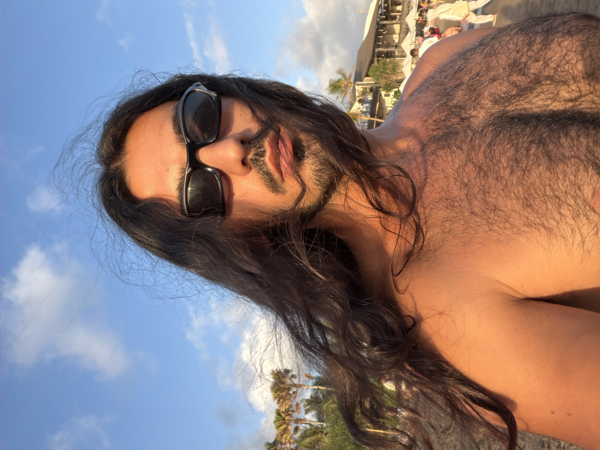 A low-angle, close-up photograph taken in North Kuta, Indonesia, depicts a man with long, dark hair and a short beard, wearing sunglasses. He is shirtless, revealing a hairy chest. The background includes a partly cloudy sky, palm trees, and a building with people near it. The lighting suggests it is daytime. The man appears to be lying down, possibly on a beach.