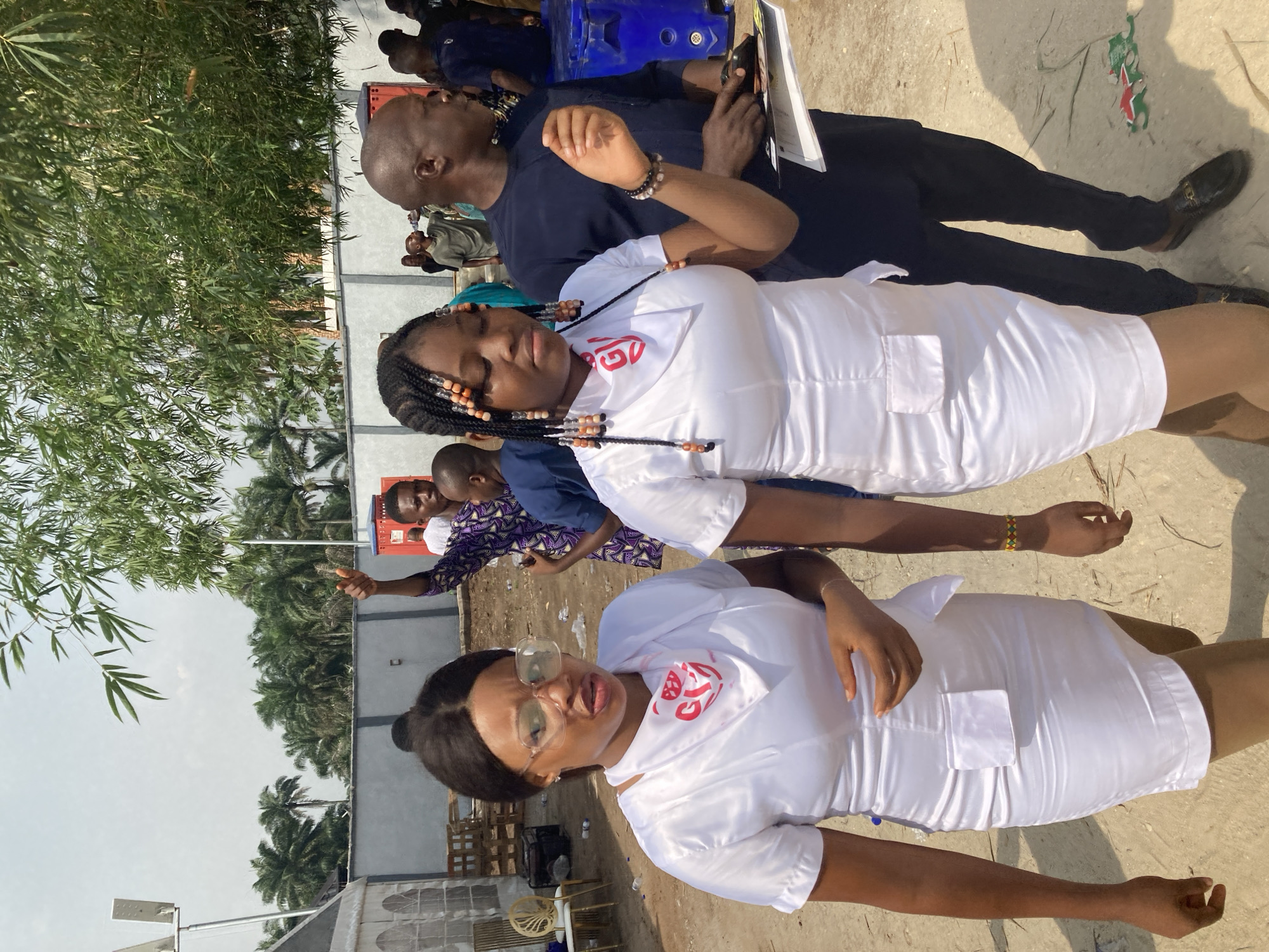 The scene depicts several individuals outdoors in Nnenu, Nigeria. Two women wearing white uniforms with red insignia stand prominently in the foreground. Behind them are other people, including a man in a dark blue shirt holding a book or tablet. There are trees and a building with a gray wall visible in the background. Some chairs and what appear to be packing crates are located in the immediate vicinity of the individuals. The ground is light-colored, possibly concrete or sandy soil.