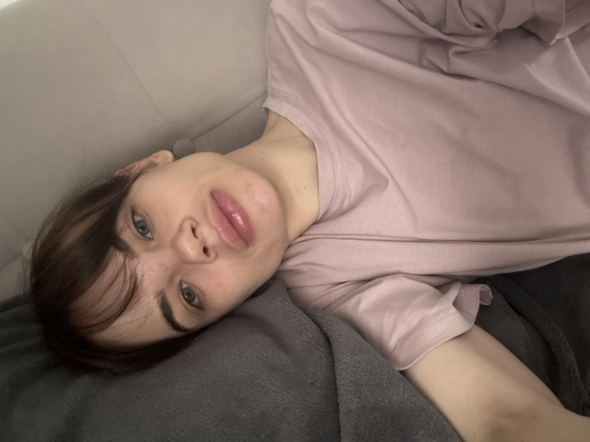A person with brown hair is lying on a gray couch or blanket, wearing a pink shirt. The focus is on the person's face, with blue eyes looking towards the camera. The background is blurred. The location is not discernable from the provided image.