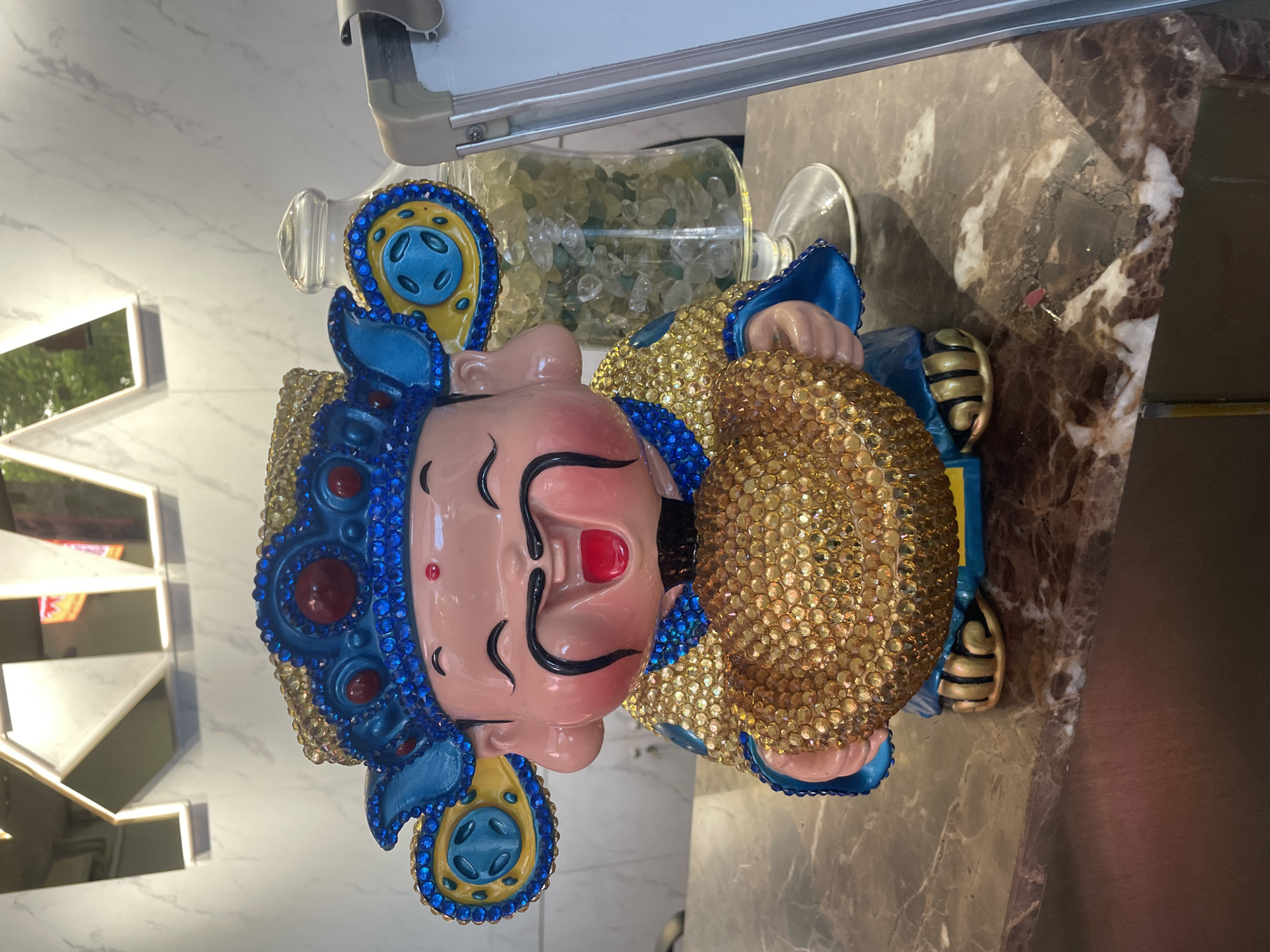 A decorative figurine of a jovial, rotund man, adorned in a blue and gold outfit encrusted with rhinestones, sits on a marble countertop. The figurine is holding a large, gold, circular object, also covered in rhinestones. The man has a red circular mark on his forehead, a black mustache, and is smiling with his mouth open. Behind the figurine, a clear glass jar filled with light green and clear stones is visible, along with the edge of a whiteboard. In the background, a large, reflective metallic "KIA" sign is mounted on a white marble-patterned wall. The scene appears to be indoors, possibly in a commercial establishment in Thủ Đức, Vietnam, suggested by the prominent KIA branding. The overall lighting is bright, illuminating the details of the figurine and its surroundings.