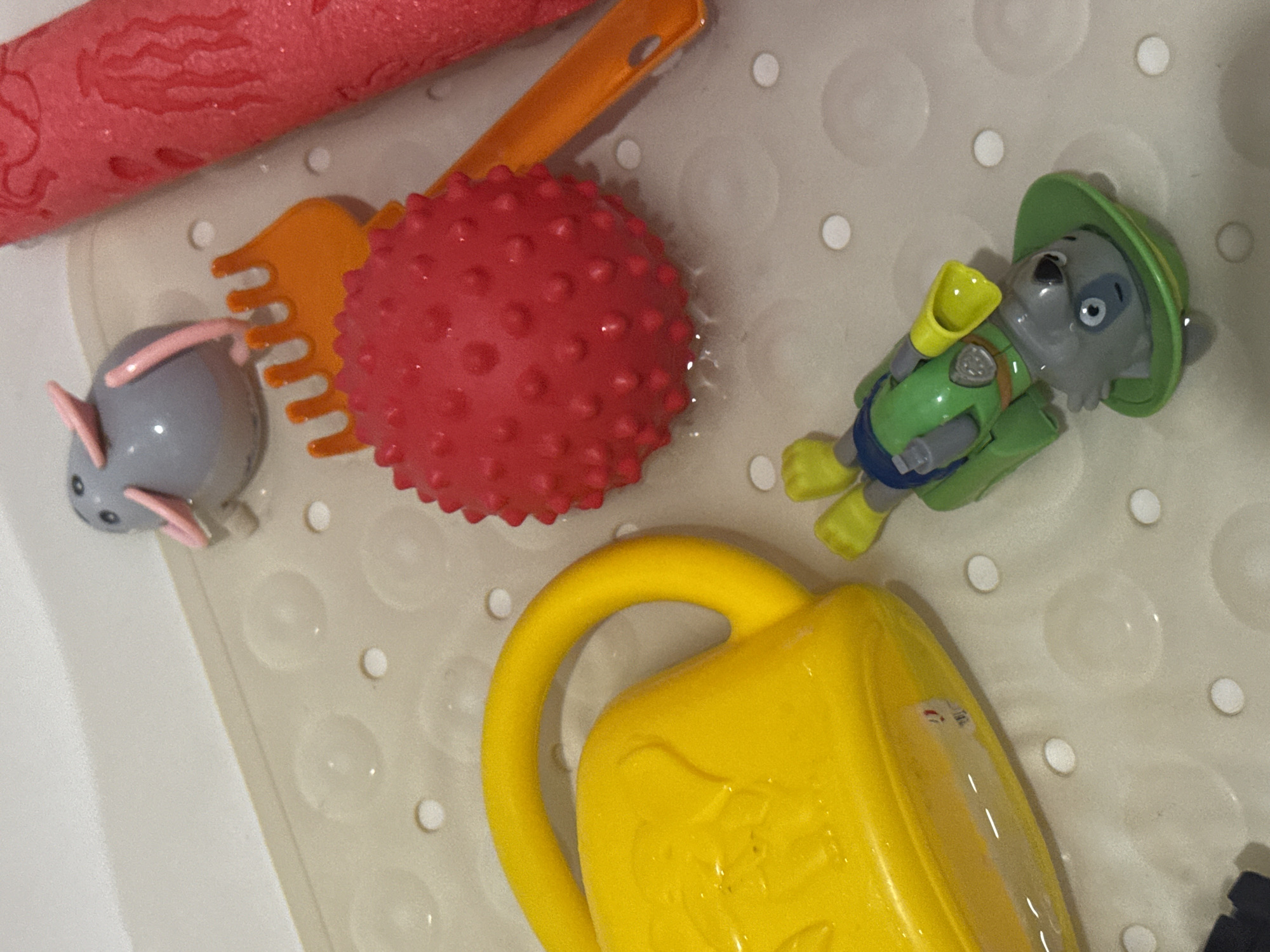 A close-up, overhead shot shows a collection of children's bath toys on a textured, light-colored surface, possibly a bath mat in a bathtub. The toys include a yellow watering can, a spiky red ball, an orange rake, a grey toy with pink ears that resembles a mouse, and a green and grey toy dog figure with yellow accents, appearing to be a character from a popular children's show. The toys are scattered loosely, and some appear to be wet, with water droplets visible on the surface and on the toys themselves, indicating recent use in water. The lighting is neutral, casting soft shadows. The scene is devoid of any human presence or other objects not typically found in a bathroom setting for children's playtime. The specific location within the city of Яромінскі сельскі Савет, Belarus, cannot be determined from the visual information provided in the image.