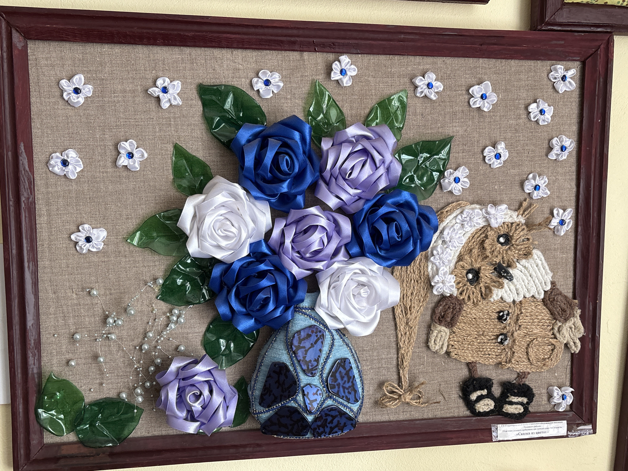This is a framed, handmade piece of art featuring a bouquet of fabric flowers and a stuffed owl figure. The bouquet, composed of satin ribbons, includes roses in shades of blue, white, and purple, arranged in a blue vase adorned with dark blue and patterned blue stones. Green leaves made of shiny material are interspersed with the flowers. Decorative strings of small, pearl-like beads add a whimsical touch. To the right of the bouquet stands a stuffed owl figure made from burlap and yarn, wearing a knitted hat and coat with button details. The owl has black button eyes and a black beak. Small white fabric flowers with blue centers are scattered throughout the composition. The artwork is presented against a tan, burlap-like background within a dark wooden frame. A small label with text in Cyrillic is visible at the bottom right of the frame. The artwork appears to be displayed on a wall. The location is stated as Ветрынскі сельскі Савет, Belarus.