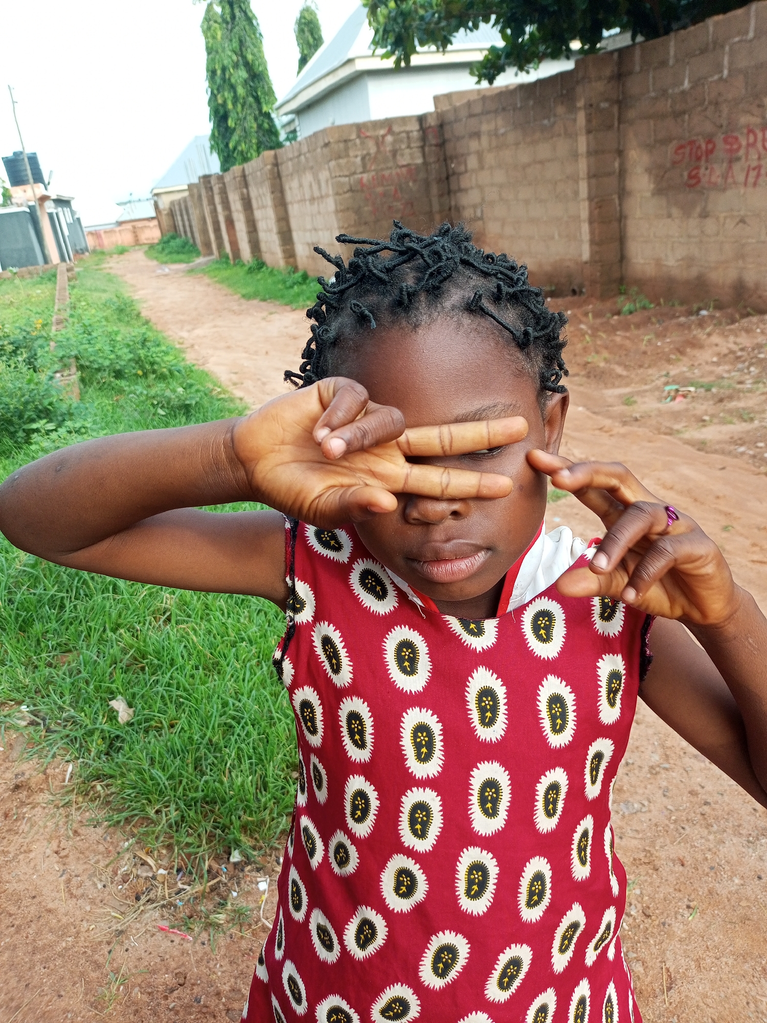 In Tempure, Nigeria, a young girl with dark skin and braided hair stands on a dirt path, her hands covering her eyes. She wears a red dress with a white and black pattern of oval shapes. The background features a brick wall with red graffiti, tall green trees, and a cloudy sky. Grass and more dirt are visible on either side of the path. The scene appears to be an outdoor residential area.