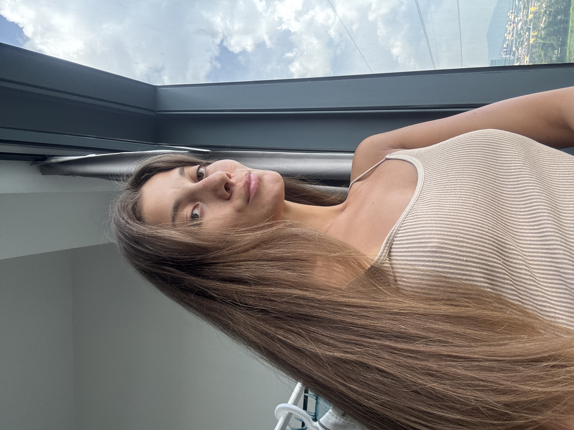 A portrait-oriented image, rotated 90 degrees counter-clockwise, features a young woman indoors. She has long, straight, light brown hair that cascades over her shoulder and arm, extending towards the bottom right of the frame. She wears a light-colored, possibly beige or cream, horizontally striped tank top with thin straps. Her head is tilted, and she looks directly at the camera with a neutral to slightly smiling expression.
The woman is positioned near a large, dark-framed window, offering an expansive view. Through the window, a bright, cloudy sky with patches of blue is visible. In the background, a cityscape with numerous buildings and distant mountains can be seen, suggesting an elevated viewpoint over an urban area. Some power lines or cables are visible across the sky. A white plastic hanger is partially visible in the bottom foreground, next to her hair. The bright lighting indicates the photo was taken during the daytime. The scene suggests a modern interior with a panoramic city view, likely in Kuala Lumpur, Malaysia. No visible text is present in the image.