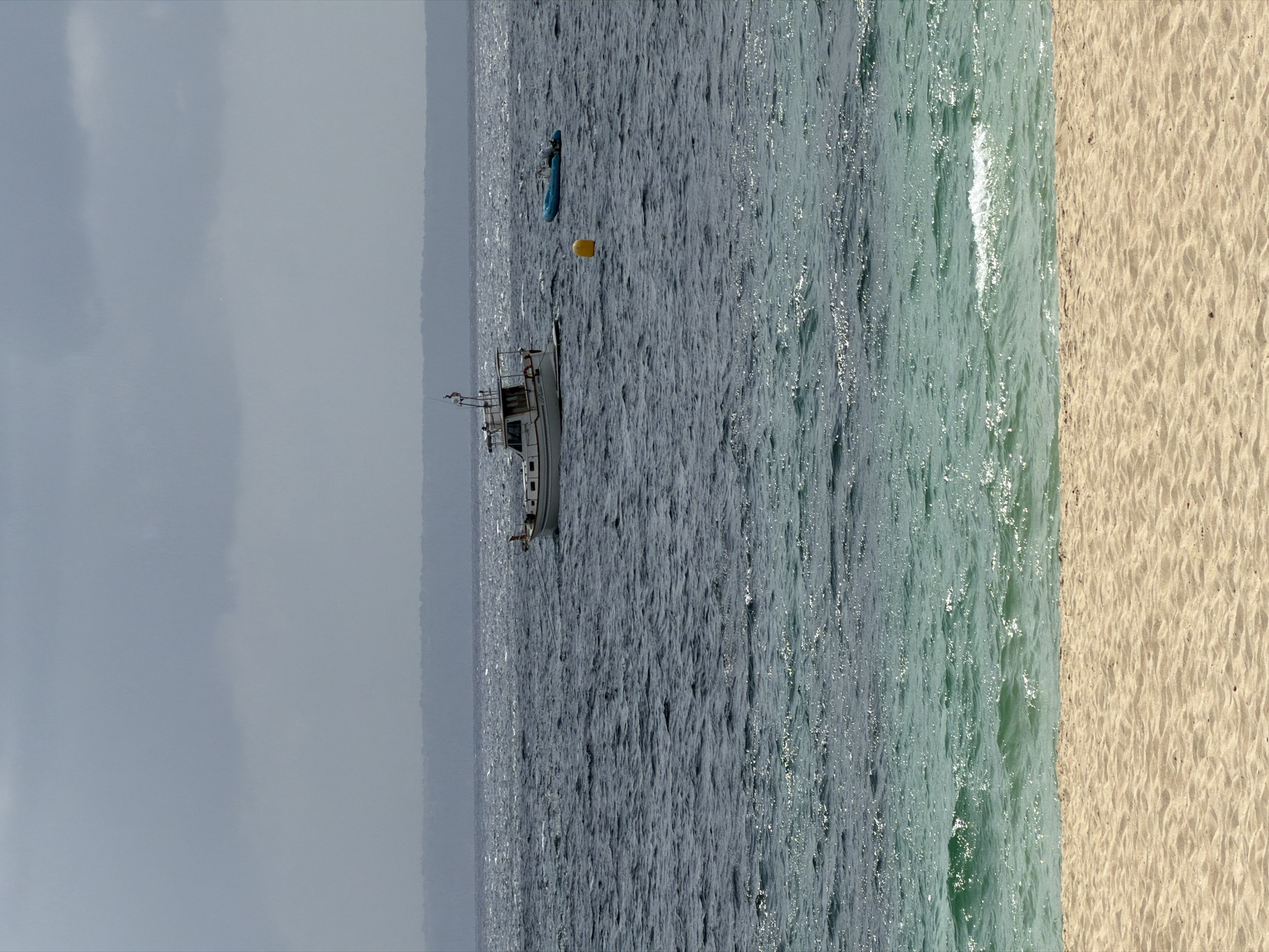 The image depicts a serene beach scene in Palmanova, Spain. In the foreground, a stretch of golden sand leads to the clear, turquoise waters of the Mediterranean Sea. The water closer to the shore is shallow and tranquil, with gentle waves lapping the sand. Further out, the sea transitions to a deeper, darker blue, with visible ripples and waves indicating a light breeze.
Dominating the middle ground of the sea are two vessels. A white fishing boat with a cabin and a small flag on its mast is anchored in the distance. To its right, a smaller blue inflatable boat with an outboard motor is also present. A yellow buoy floats between the two boats. In the far distance, a hazy silhouette of land is visible, likely the coastline of Mallorca. The sky above is overcast with a soft, diffused light. The overall impression is one of a calm day at the beach, with a sense of quietude and natural beauty.