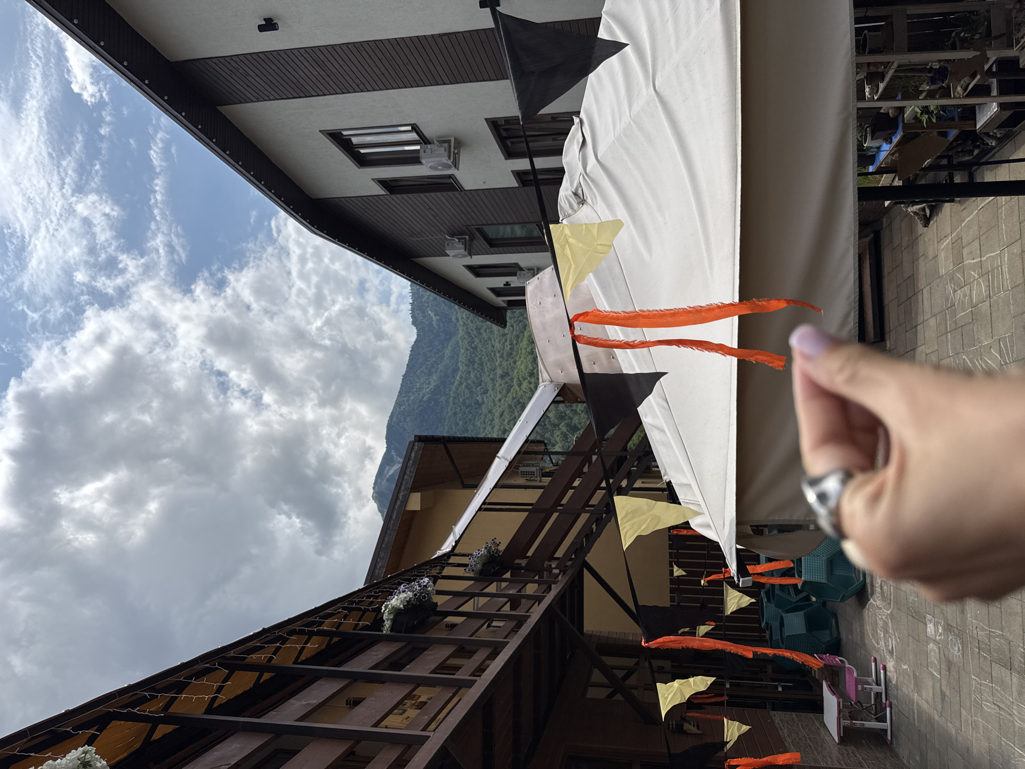 The image depicts an outdoor scene with buildings and a mountain range in the background, likely in Krasnaya Polyana, Russia, known for its mountain resort environment. In the foreground, a hand with light purple nail polish and a silver ring is held up, partially obscuring the view. Hanging in the air are colorful pennant banners, primarily black, yellow, and orange, strung across what appears to be a covered outdoor seating area or market stall.
To the left, a multi-story wooden building with balconies and hanging flower baskets is visible. Adjacent to it is a lighter-colored building with dark wood siding and visible air conditioning units. The ground is paved with square tiles, and some chalk drawings are faintly visible on the pavement. In the mid-ground, there are several green, cube-shaped chairs and a small pink and white table with chairs, suggesting a child's play area or outdoor dining setup. The sky is partly cloudy with patches of blue. The overall impression is of a resort or guesthouse area, possibly decorated for a festive occasion.