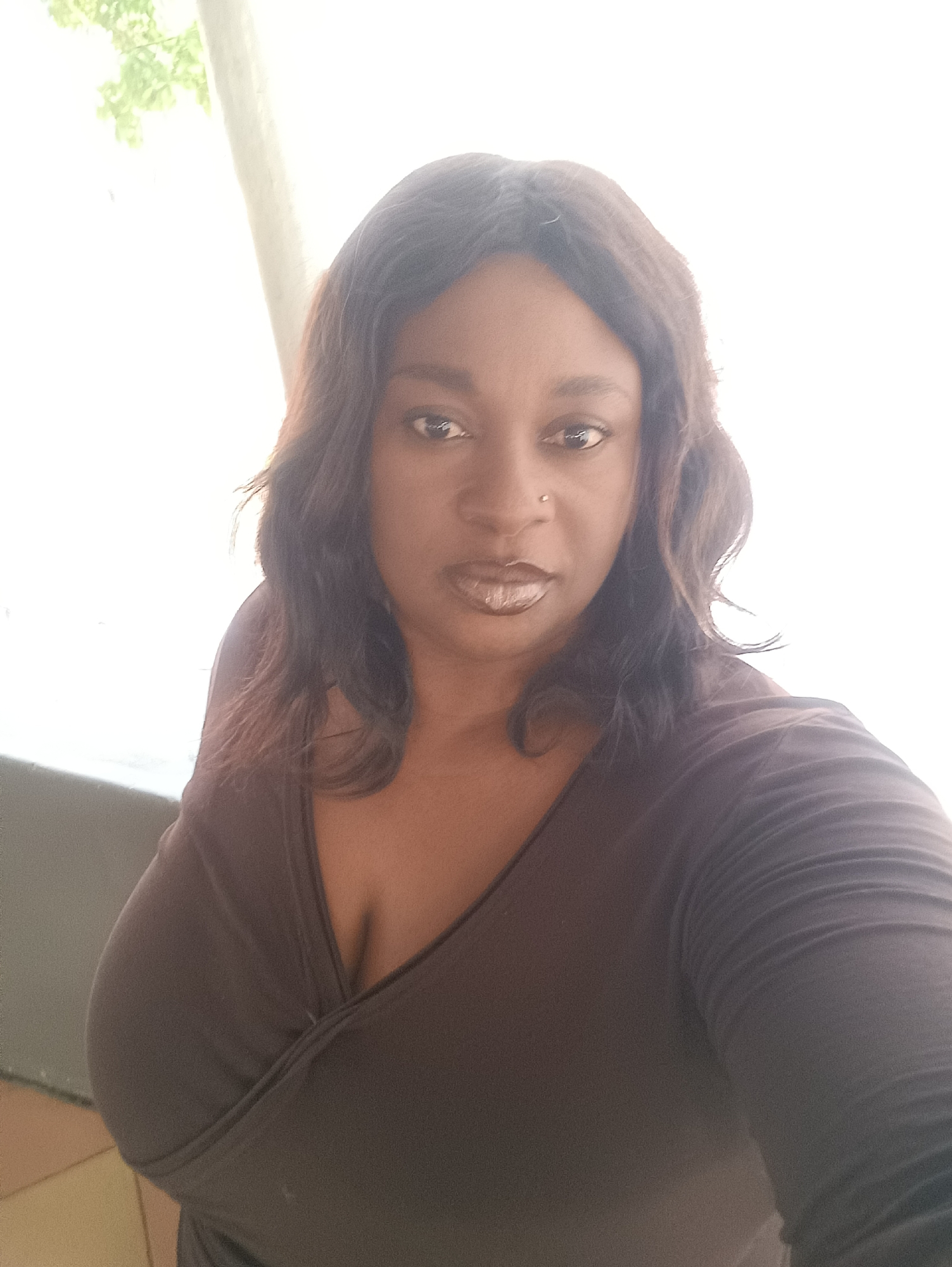 A medium shot depicts a dark-skinned woman wearing a dark, long-sleeved shirt with a V-neck. She has shoulder-length dark hair with auburn highlights. Her features include a nose piercing, dark eyes, and metallic lipstick. The background is bright and appears to show a window with green leaves visible outside, suggesting an indoor or semi-indoor location. The scene's exact location within Jos, Nigeria, is not discernible from the image.