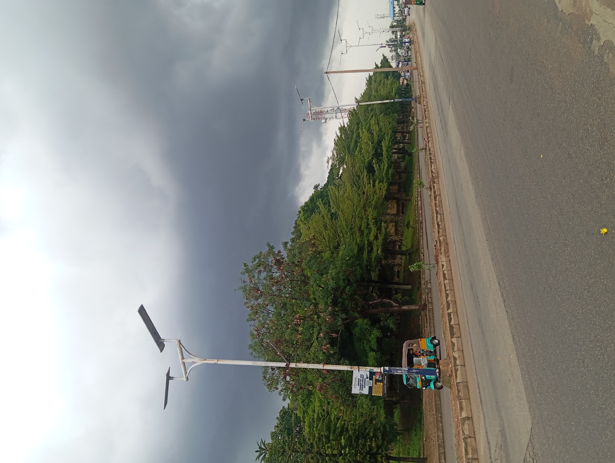 An overcast, gloomy day is depicted on a street in an unknown location. The sky is a deep, dark gray, filled with heavy clouds, suggesting an impending storm. The street is asphalt and appears to be a multi-lane road. On the left side of the road, a median separates it from a sidewalk and a grassy area lined with trees. A tall lamppost with two solar panels stands prominently on the left. A sign is attached to the lamppost. A three-wheeled auto-rickshaw, a common mode of transport in some regions, is stopped near the lamppost, with people visible inside. Further down the road, there are more lampposts and trees lining the street. In the distance, a cell tower is visible, and other vehicles are present on the road. The overall atmosphere is subdued due to the weather conditions.