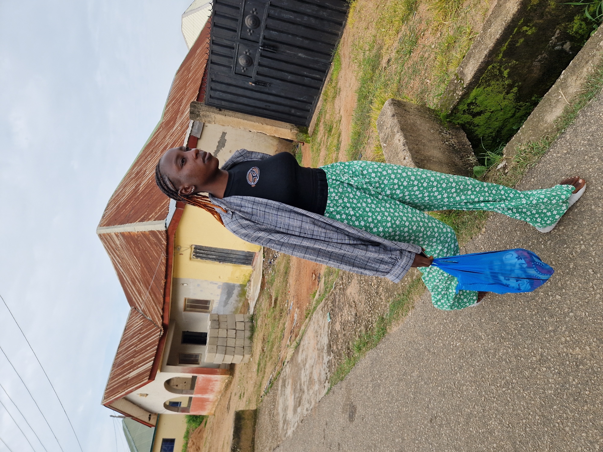 A young Black female walks on a paved path next to a drainage ditch in Kabong, Nigeria. She is wearing a black t-shirt, a plaid open shirt, and green patterned pants. In her right hand, she carries a blue plastic bag. In the background are several single-story buildings with corrugated metal roofs, some appearing unfinished, and a large black metal gate. The sky is overcast.