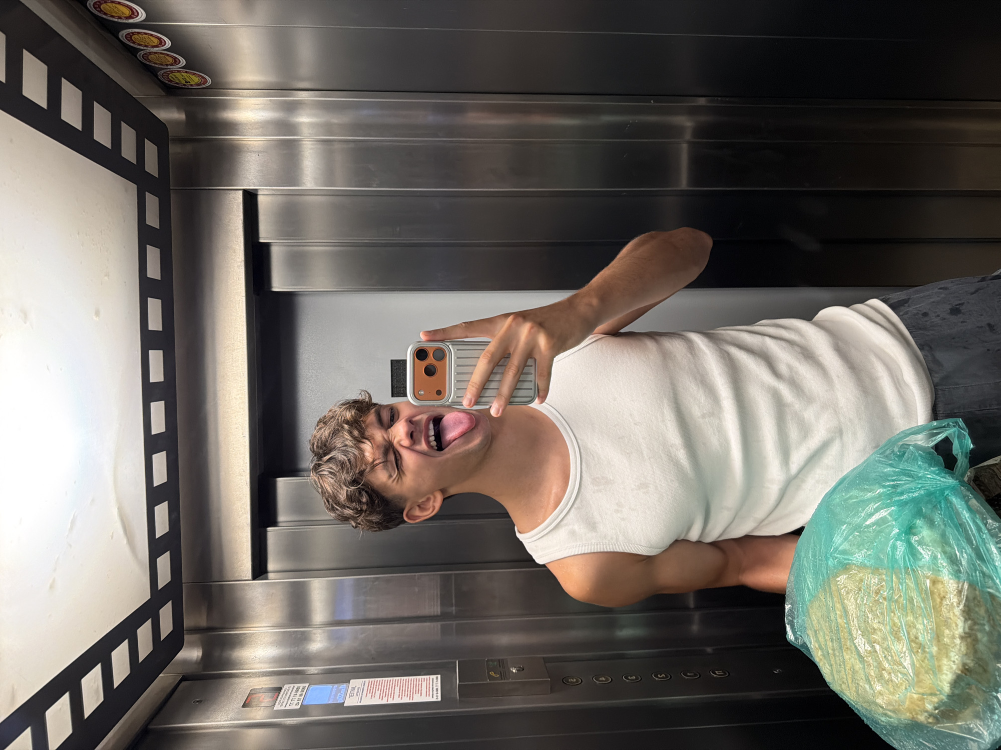A young man is taking a selfie in an elevator. He is wearing a white tank top and dark pants, and he is holding a large, round object wrapped in green plastic. His tongue is sticking out, and he is winking at the camera. The elevator walls are made of brushed metal, and there are buttons and signs on the wall. The ceiling of the elevator is white with black decorative squares. The image was taken in an elevator in Blagoevgrad, Bulgaria.