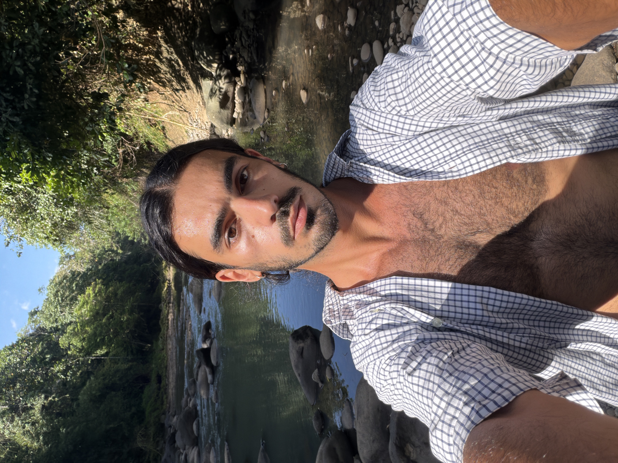 A man with dark hair and a beard is outdoors, next to a river or stream. He is wearing an unbuttoned, checkered shirt. The surrounding environment features lush green vegetation and rocks along the riverbank. The location is in a natural setting, specifically in Selemadeg Timur, Indonesia.