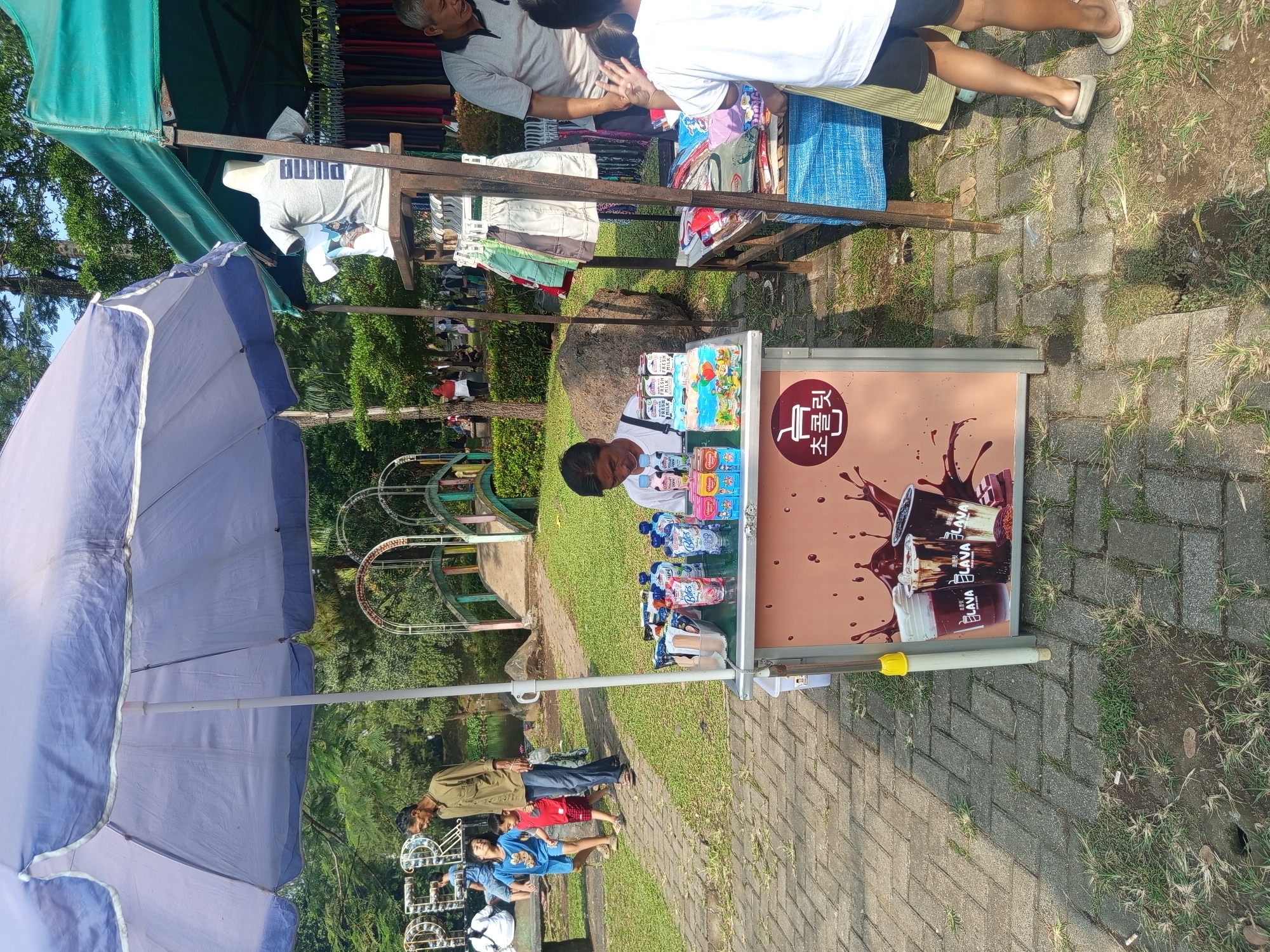 In the city of Ciomas, Indonesia, a scene unfolds at a park. A man, presumably a vendor, sits behind a stall filled with beverages. In front of him, on a table, are numerous small bottles and cartons of drinks, including what appears to be milk and juice. The stall itself is decorated with an image of three iced drinks. To the left, a large, light blue umbrella provides shade. Beyond the stall, a pathway made of interlocking bricks leads through a grassy area with trees and a body of water. In the background, a decorative bridge with arched structures can be seen. To the right of the stall, another vendor's setup is visible, featuring a rack with clothing items, including t-shirts and shorts, and a table with fabric. Several people are present in the park, including a man in a tan shirt walking with two children, and a woman in a red headscarf in the distance. The overall impression is a lively outdoor market or gathering.