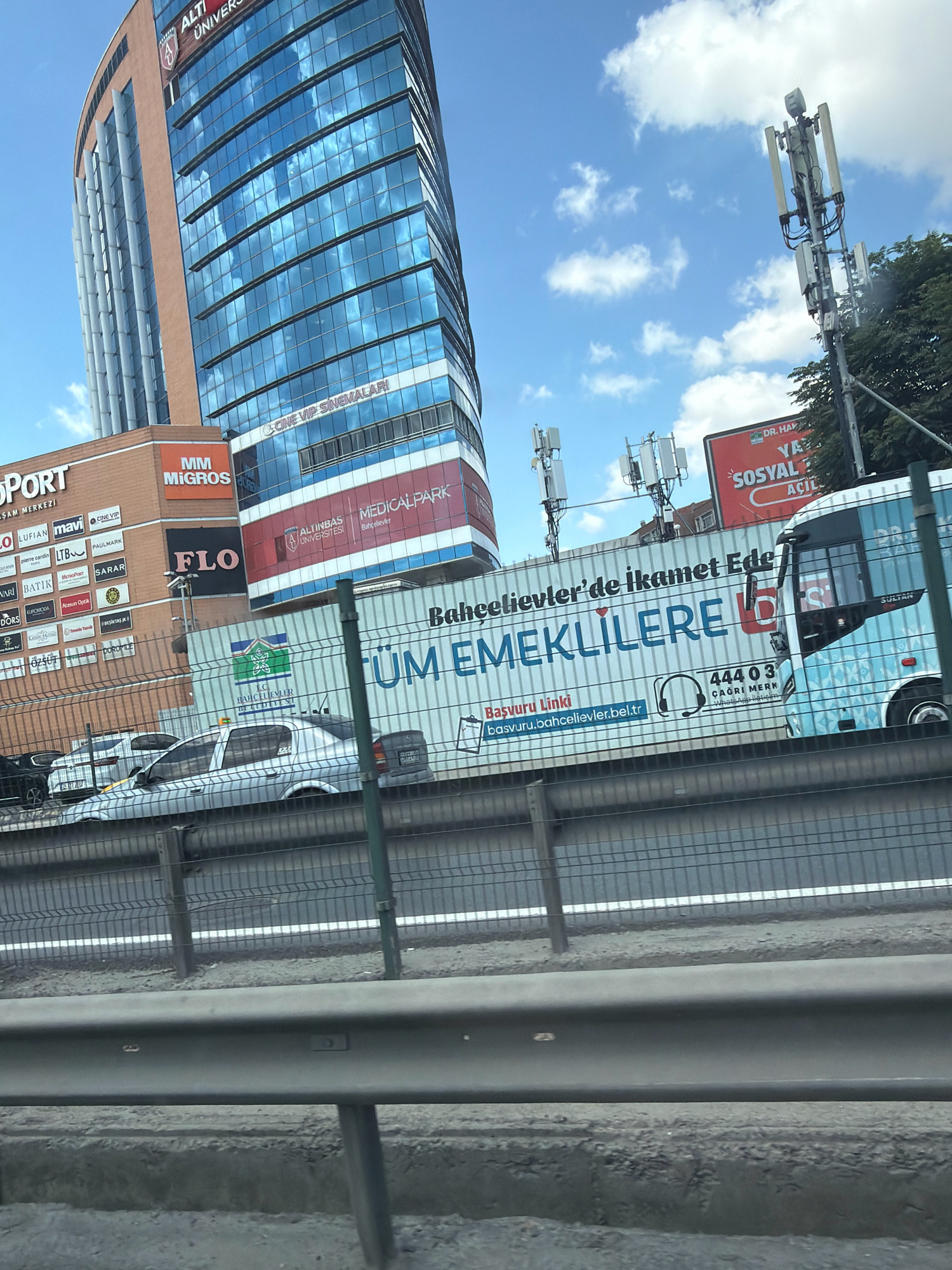 The image shows a street scene in Bakırköy, Turkey, with a tall modern glass-fronted building dominating the background. The building has a curved facade and is adorned with reflective panels that mirror the sky and clouds. Several businesses are advertised on the building's lower levels, including "ALTINBAŞ UNIVERSITY," "MEDICAL PARK," "CINE VIP SINEMALARI," and "MİGROS." Adjacent to the tall building is a lower brick-faced building with more commercial signage.
In the midground, there is a white truck or container with large blue text that reads, "Bahçeşehir'de ikamet Ede... / TÜM EMEKLİLERE D..." which translates to "To reside in Bahçeşehir... / For all retirees..." and includes a phone number and website. A portion of a light blue bus is also visible on the right side of the image. Several mobile phone towers are positioned on top of buildings.
In the foreground, a highway or road is visible with a metal safety barrier. Cars are seen on the road, including a silver sedan. A chain-link fence runs along the edge of the road, separating it from the area where the buildings are located. The sky is partly cloudy and a bright blue. The overall impression is an urban commercial area with a mix of modern and older architecture.