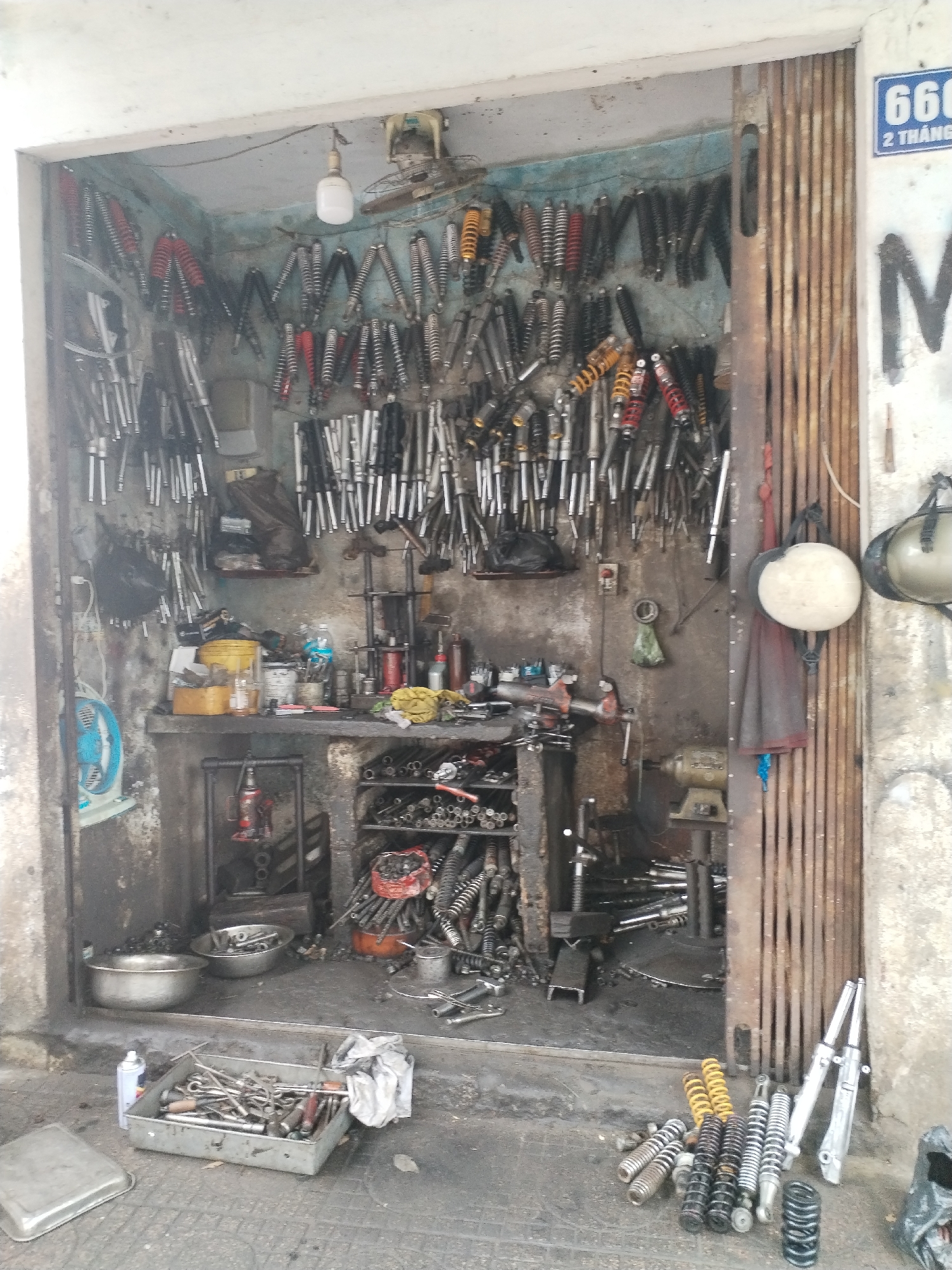 The image shows a cluttered roadside motorcycle repair shop in Nha Trang Ward, Vietnam. The shop is small and densely packed with motorcycle parts, particularly shock absorbers, which are hung on the back wall and stacked on shelves. Various tools and equipment are scattered throughout the space, including wrenches, metal basins, and a hydraulic jack. The shop has a partially open metal gate, and motorcycle helmets are hung on it. The overall impression is of a busy, well-used workspace.