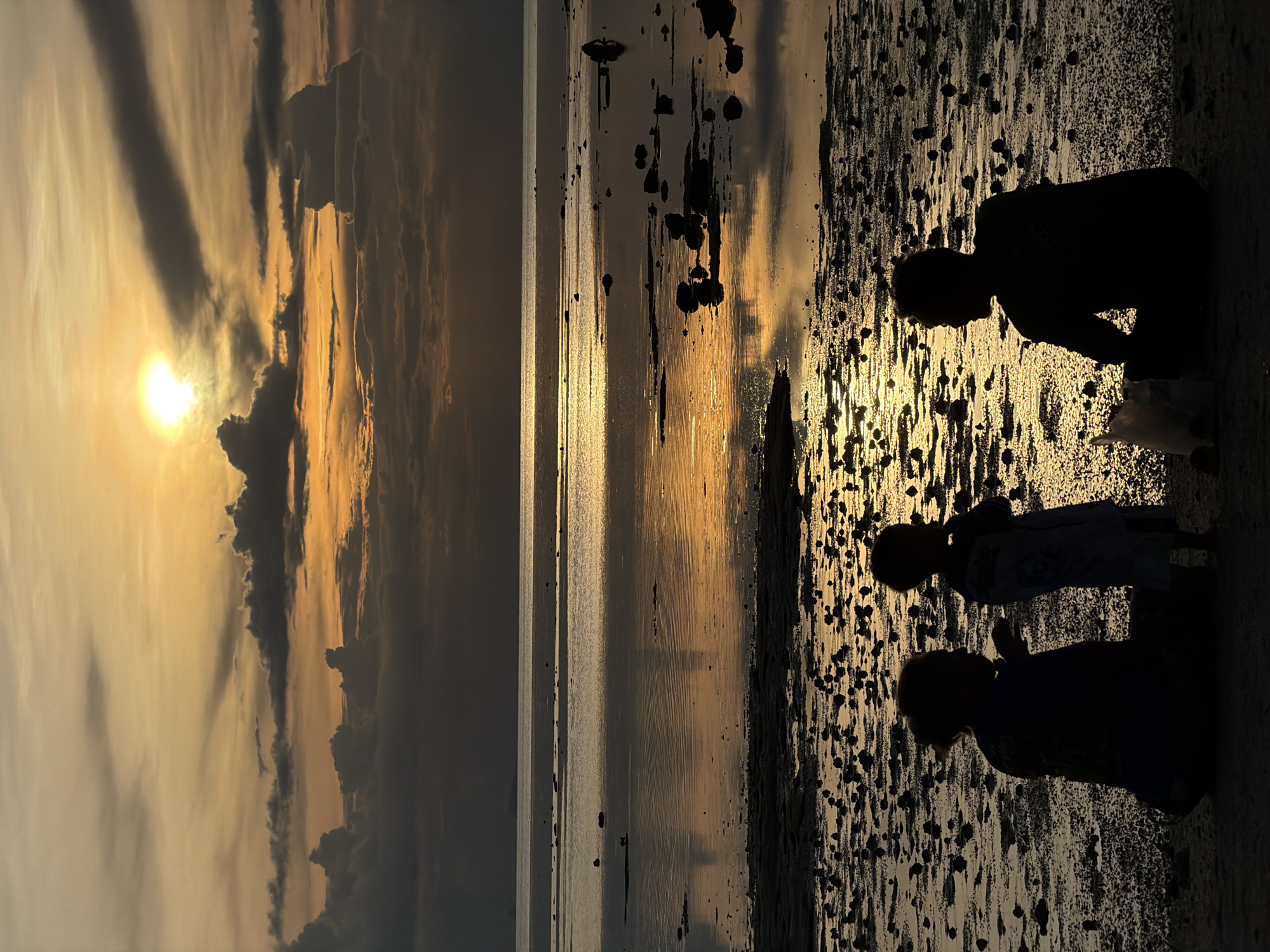 The image depicts a sunset at Baan Plai Laem, Thailand. In the foreground, silhouetted against the warm glow of the setting sun, are three figures sitting on the sandy shore. Two of the figures, one adult and one child, are facing towards the sea, while the third figure, an adult, is seated with their back to the viewer, also facing the water. They are positioned on a beach scattered with small rocks and tide pools that reflect the dramatic sky. The ocean stretches out to the horizon, with a gentle swell and the sun, partially obscured by clouds, casting a golden light across its surface. The sky is filled with a mix of dark, heavy clouds and bright, luminous patches, creating a striking contrast. The overall atmosphere is serene and contemplative, capturing a moment of quiet observation of the natural world.