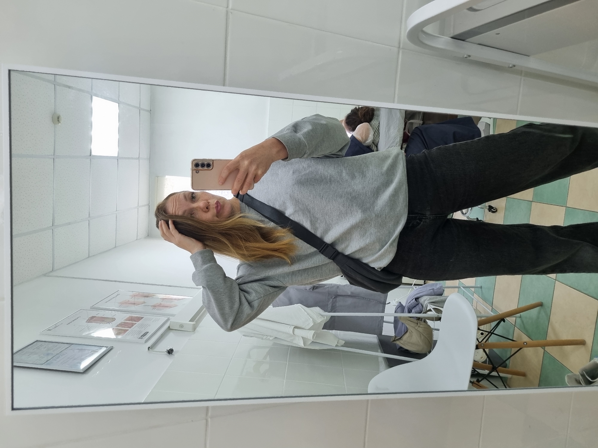 A woman is taking a mirror selfie in what appears to be a medical examination room. She is wearing a gray sweatshirt and dark jeans, with a black cross-body bag. Her right hand is in her hair, and she is looking at her phone, which is held in her left hand. In the reflection of the mirror, another person is lying on an examination table with their head resting on a pillow. The room has tiled walls and a checkered floor. There is a white chair to the left of the frame, and several items of clothing are hanging on a rack in the background. A framed document is visible on the wall. The overall impression is a casual snapshot taken in a clinical setting.