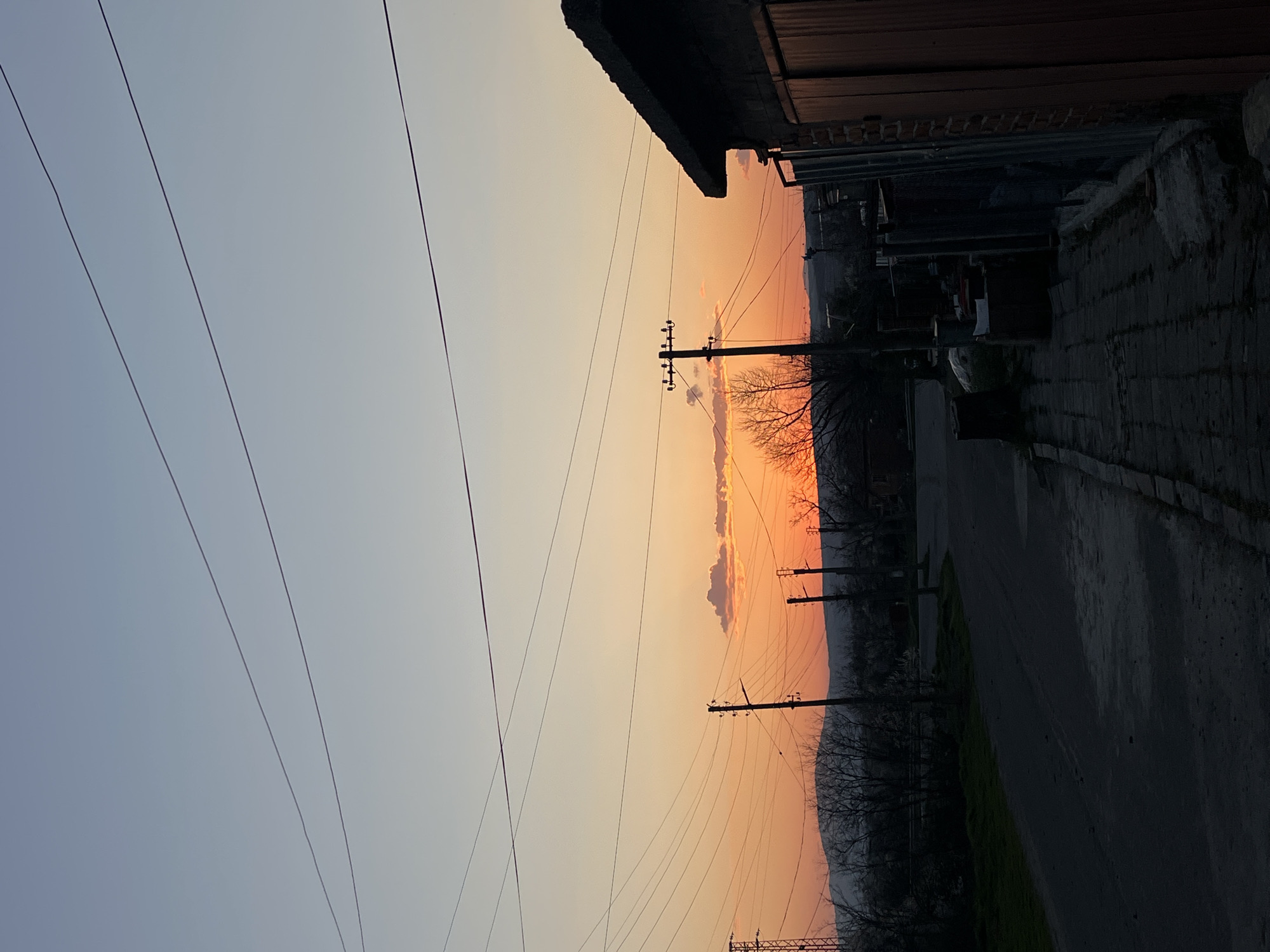 The image captures a street scene in Lozarevo, Bulgaria, at sunset. Power lines are visible against the colorful sky. Utility poles stand along the street. A building lines the right side of the frame, with a sidewalk and street running adjacent to it. The sky is tinted with orange and yellow hues. A narrow, tree-lined ridge is visible in the distance. A vehicle is parked next to the building.