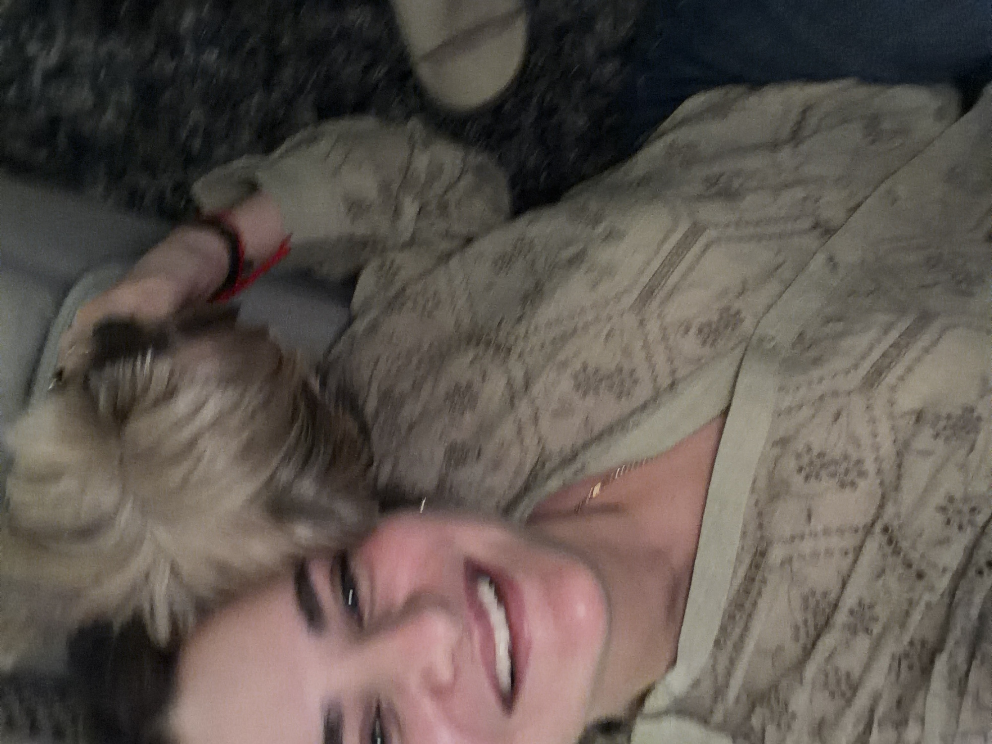 A woman with blonde hair pulled back in a bun is lying down and smiling at the camera. She is wearing a tan patterned shirt and a gold necklace. A red and black bracelet is visible on her wrist. She is lying on a grey surface. A sandal is partially visible in the upper portion of the image. The location is unknown.