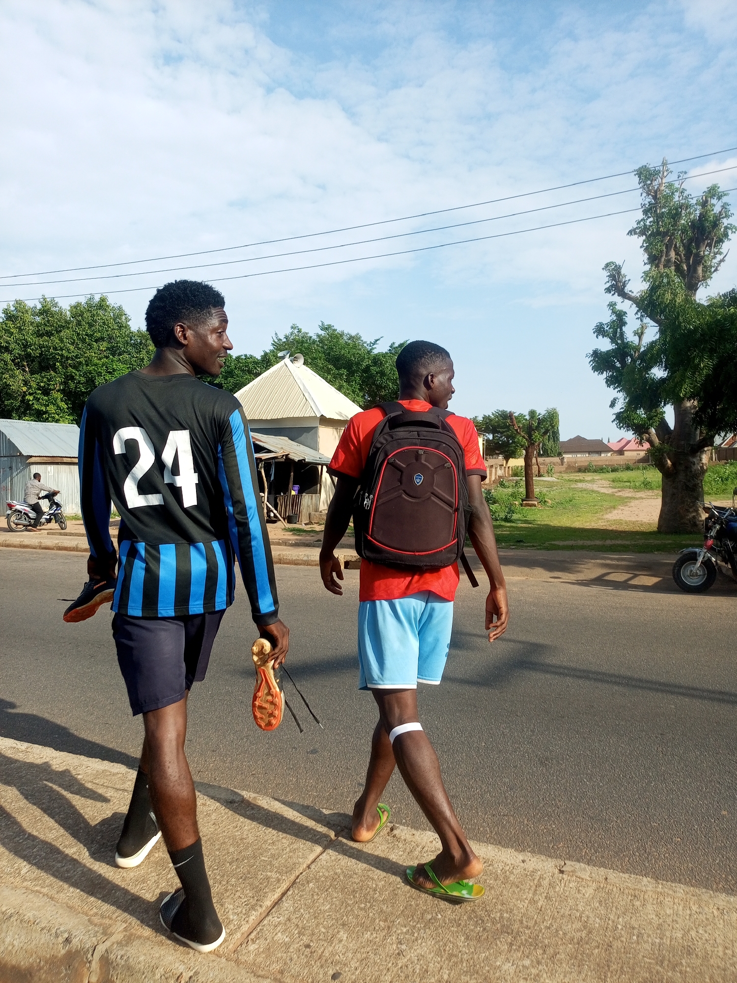 Two young men walk along a paved road, with a sidewalk on their left. The man on the left is wearing a long-sleeved black shirt with blue stripes and the number "24" in white on the back. He is also wearing dark blue shorts and black sneakers with white soles. He is carrying a pair of soccer cleats in his right hand. The man on the right is wearing a red t-shirt, light blue shorts, and green flip-flops. He is carrying a black backpack with red trim on his back. The sun is high in the sky, casting shadows on the ground, suggesting it is daytime, likely late morning or afternoon. The weather appears clear with a few scattered clouds. Behind the men, there are trees and some buildings, including what appears to be a small market stall. In the background, a person is riding a motorcycle on the road. The overall scene suggests a casual stroll on a warm day in a somewhat rural or suburban area of Gombe, Nigeria.