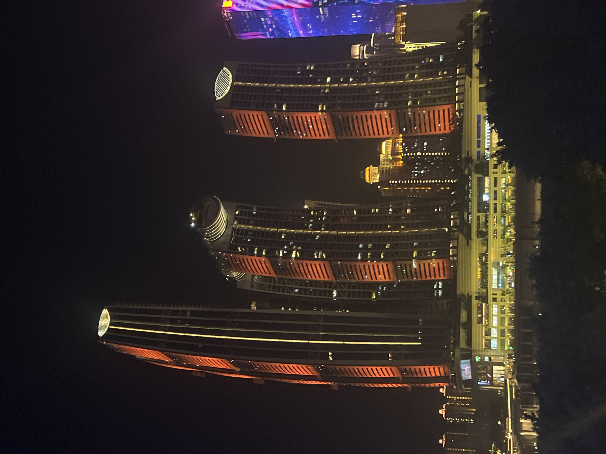 In the heart of Jiangbeicheng Subdistrict, China, a nighttime cityscape unfolds, dominated by towering skyscrapers illuminated by a vibrant spectrum of lights. Three prominent skyscrapers stand as the focal point, each with a unique architectural design. The skyscraper on the left is exceptionally tall and slender, featuring a distinctive oval-shaped illuminated top. Its facade is a dark grey, punctuated by vertical golden lights and sections of bright orange lighting that wrap around the building in staggered horizontal bands.
To its right, a cluster of three skyscrapers share a similar architectural style, with angled facades and segmented orange lighting. The middle skyscraper in this cluster has a curved top, reminiscent of a large dome. These structures are juxtaposed against the night sky, which is almost completely black, allowing the artificial lights to cast a dramatic glow.
Further in the background, other buildings of varying heights and designs can be seen. Some exhibit a more traditional rectangular shape, while others are adorned with a soft, warm yellow light. One building in the far right displays a striking vertical pattern of pink and blue lights, adding a touch of modernity to the scene.
At the base of the central cluster of skyscrapers, a multi-story commercial building is visible, characterized by a horizontal design and illuminated windows. The lower levels of this building are a pale yellow, suggesting a shopping mall or office complex. The entire scene is framed by a dark foreground of trees, their silhouettes stark against the illuminated structures. The reflection of some lights can be seen on what appears to be a body of water in the lower left corner of the image. The overall impression is one of a bustling, modern urban environment alive with nocturnal activity and architectural splendor.