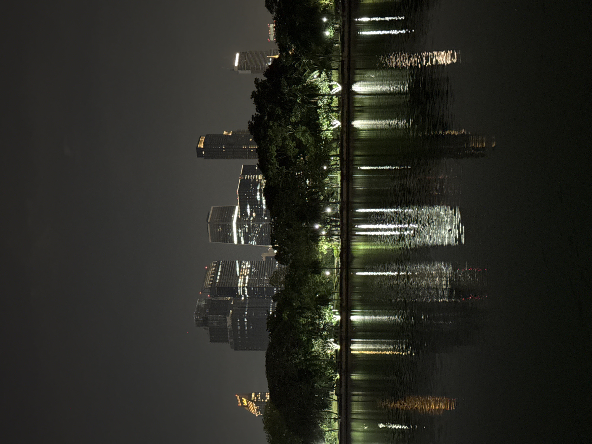 The photograph captures a nighttime scene in Bangkok, Thailand, featuring the city skyline reflected in a body of water. Buildings, illuminated with lights, stand tall against the night sky, their reflections creating distorted, shimmering patterns on the water's surface. A line of trees runs parallel to the water's edge, their dark forms punctuated by occasional streetlights that add to the overall luminosity of the scene. The night sky is a uniform dark gray.