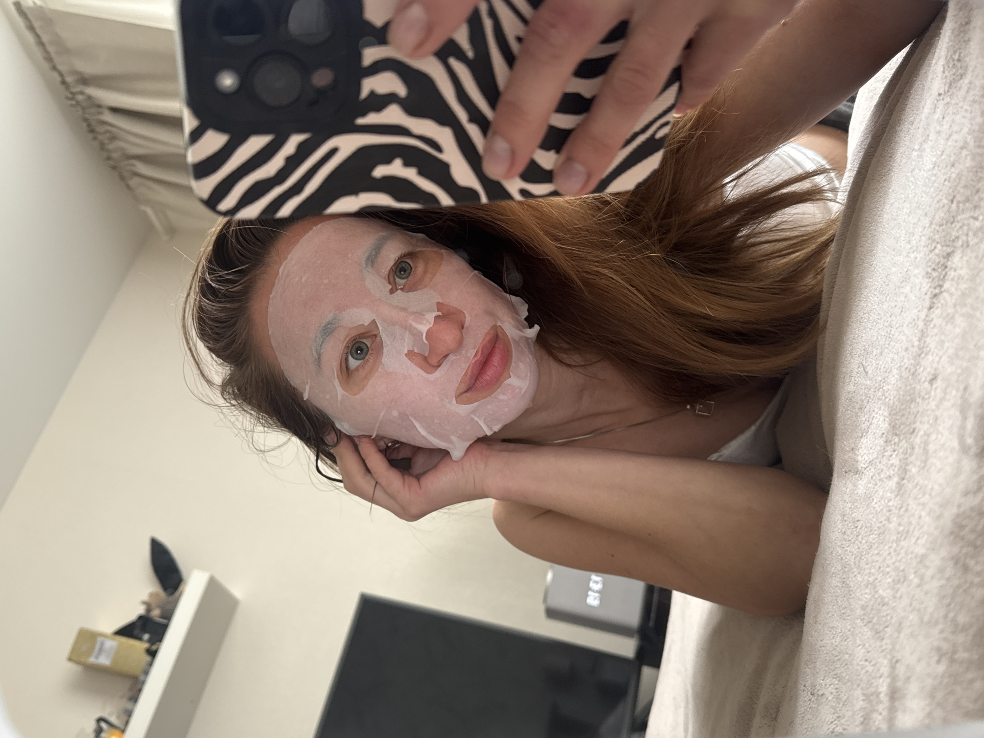 The image displays a woman engaged in a self-care routine, taking a selfie while wearing a white sheet face mask. She has long, reddish-brown hair and striking green eyes, looking directly at the camera with a calm expression. Her right hand, with neatly kept nails, holds a smartphone featuring a distinctive black and white zebra print case, which dominates the upper part of the frame. A delicate silver necklace with a small square pendant is visible around her neck.
The setting is an indoor room, likely a bedroom or living area, suggested by the light-colored, textured blanket or bedding she is reclining on. In the background, a plain light-colored wall is visible, on which a white floating shelf is mounted. The shelf holds several items, including a small cardboard box and a dark, indistinct object. Below the shelf, a section of a dark flat-screen television is visible. To the right of the TV, a grey electronic device, possibly a digital clock or smart speaker, displays what appears to be "BE EX" when rotated, which likely translates to "18:13" or "13:18," indicating the time of day is either early evening or early afternoon. The overall composition suggests a moment of relaxation. The image is captured from a slightly tilted perspective. The scene takes place in Chaikovsky, Russia.