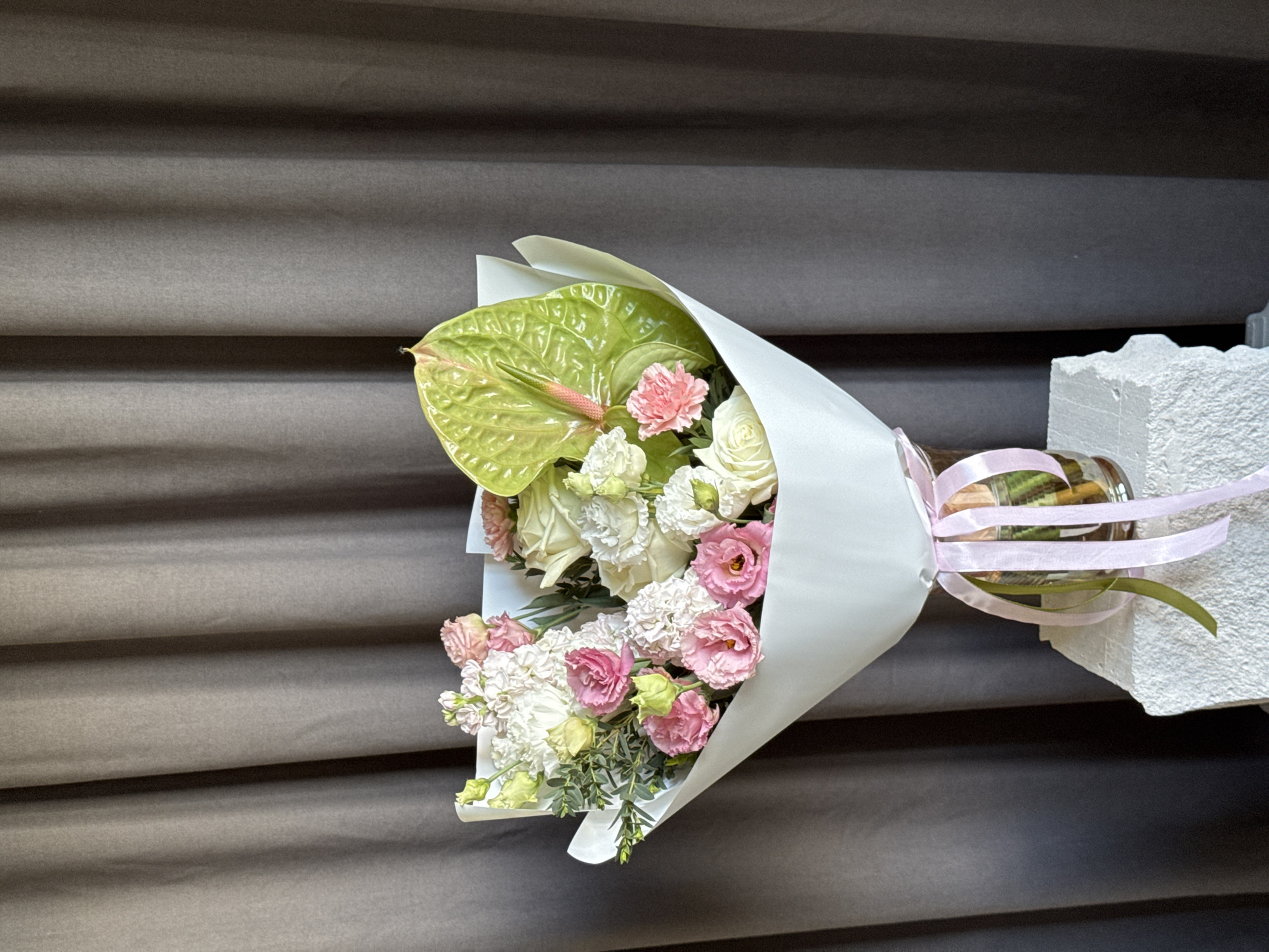 A bouquet of fresh flowers, predominantly white and pink, is centered in the frame against a backdrop of dark grey-brown fabric curtains. The bouquet is wrapped in plain white paper, forming a cone shape.
The floral arrangement includes a prominent large, glossy, heart-shaped green Anthurium leaf with a pink spadix, positioned towards the top right. Other flowers visible include ruffled pink Lisianthus (Eustoma) blooms, numerous white Stock (Matthiola) spikes with multiple small florets, creamy white roses or large Lisianthus, and smaller pink carnations. Green foliage fillers are interspersed throughout the arrangement.
The bouquet is placed in a clear glass cylindrical vase, which is visible below the white wrapping. The vase contains water and the lower stems of the flowers. Around the neck of the vase, multiple satin ribbons in light pink and olive green are tied in a bow, with the ends cascading downwards.
The vase rests on a textured, light-colored, possibly concrete or rough plaster block, which appears irregularly shaped on its left side. The background consists of vertically draped curtains, exhibiting soft folds and a matte texture, uniformly dark grey-brown in color. The lighting is even and appears to originate from the front, illuminating the bouquet and creating soft shadows.