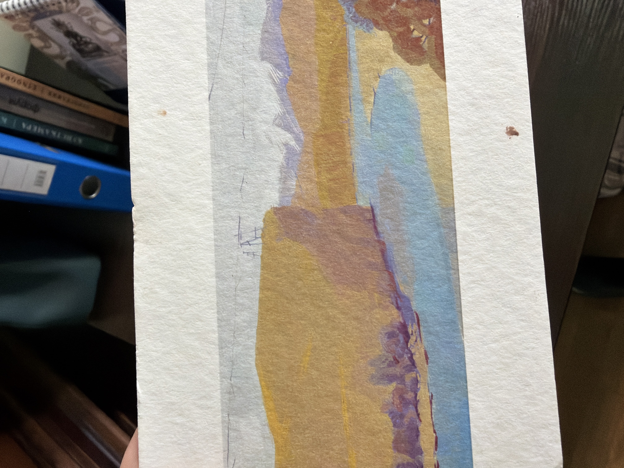 The image shows a partially visible watercolor painting. The style is impressionistic, with broad strokes of color. The painting features a vertical composition with colors including yellow, blue, lavender, and brown. It seems to depict a landscape with vertical architectural elements or rock formations. The painting is on white watercolor paper and is held in front of a bookshelf with books and a blue file folder. The painting appears to be an interpretation of a scene, possibly inspired by Saint Petersburg's architecture or natural landscape, but it lacks specific, identifiable landmarks to confirm a precise location within the city.