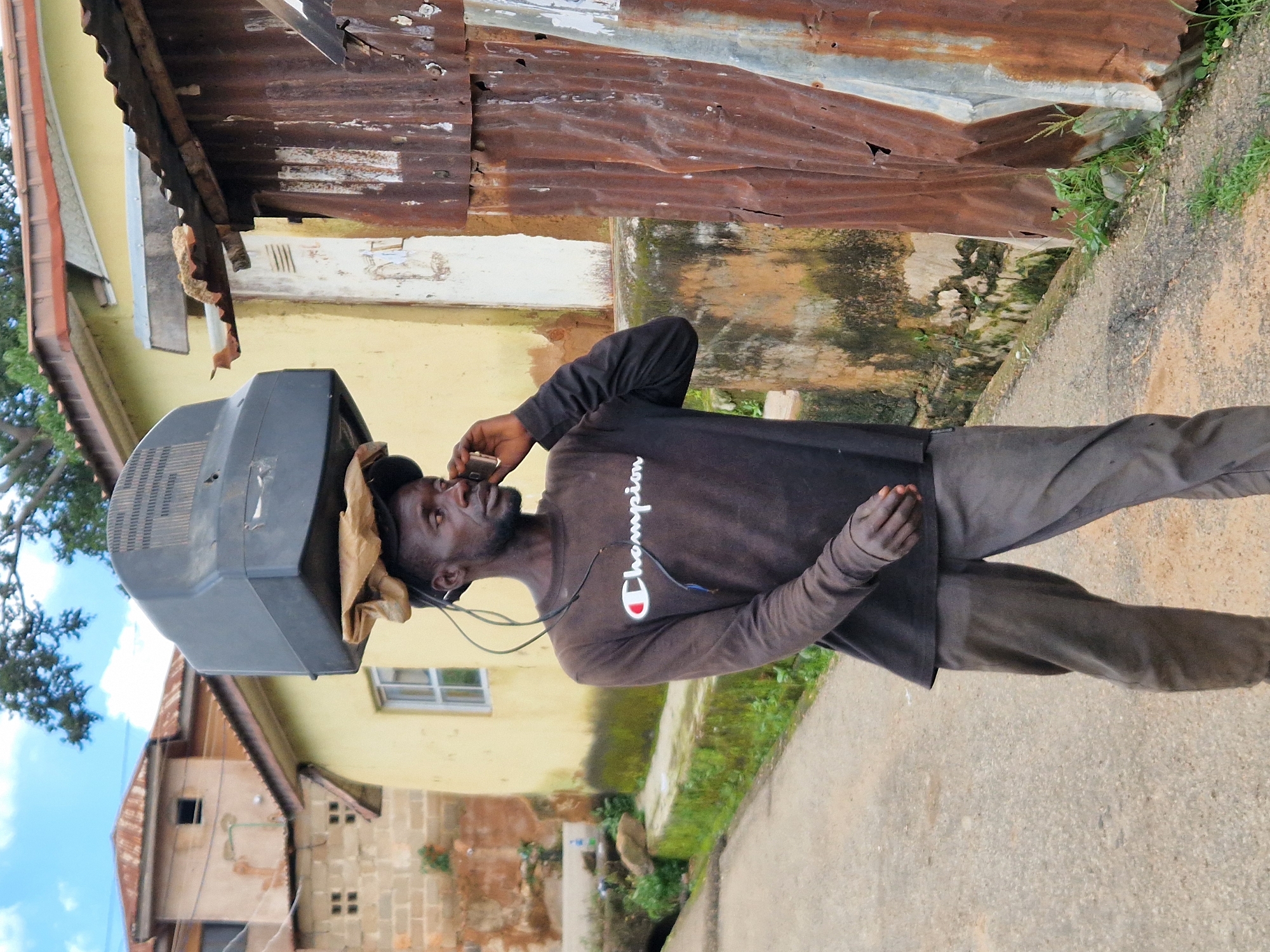 A Black man walks along a paved street, carrying a television on his head. He wears a dark long-sleeved Champion shirt and dark pants. He is talking on a cell phone held to his right ear. Wires from the television hang down around his head and neck. The man is in an outdoor urban environment. To his left is a yellow building with a red-tiled roof and a window. To his right are two corrugated metal structures, one heavily rusted and the other lighter in color. Green vegetation is visible along the side of the street and against the buildings. The sky is partly cloudy.