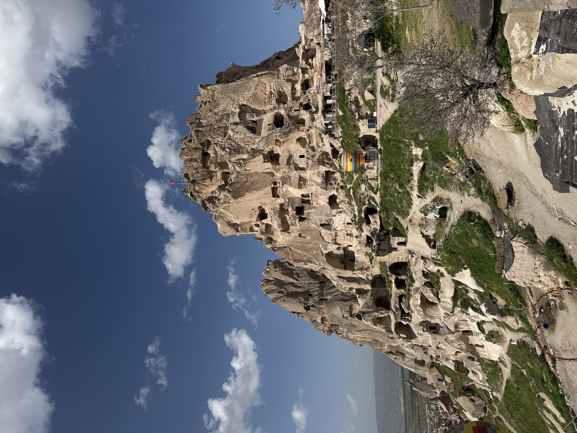 The image depicts Uçhisar Castle, a large rock formation riddled with caves and dwellings, located in Uçhisar Beldesi, Turkey. The structure is primarily composed of tan-colored rock. Various openings of different sizes, indicating former or current cave dwellings, are visible across its facade. A Turkish flag is positioned atop the highest point of the castle. Vegetation, including grass and a bare tree, grows on the ground around the castle. A partially visible hot air balloon is near the base of the castle. The sky above features a bright blue color, punctuated by scattered white clouds. In the background, there is a view of the surrounding valley.