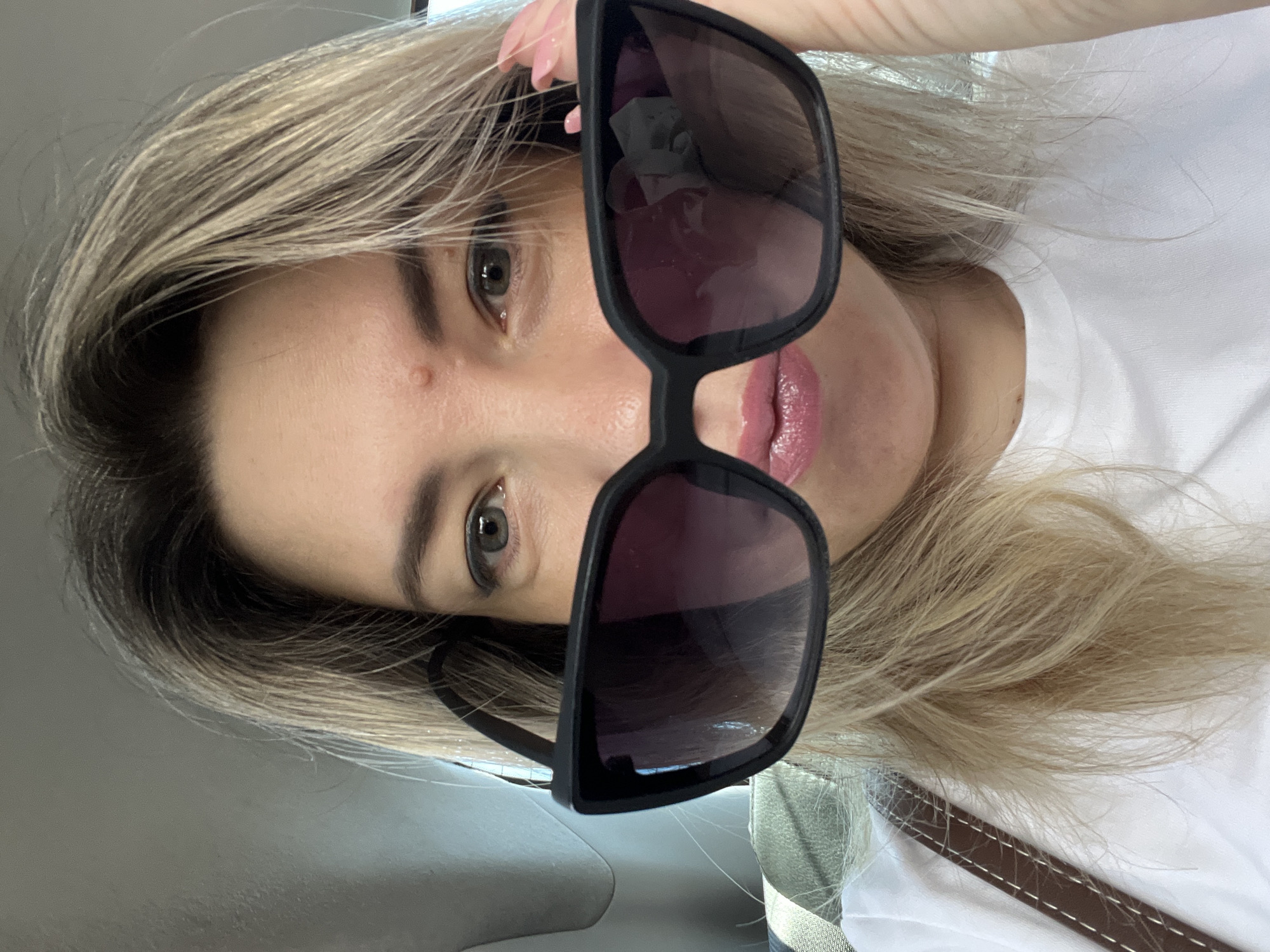This is a close-up, front-facing selfie of a woman indoors, possibly in a vehicle. She is holding large, black-rimmed sunglasses up to her face, obscuring the upper portion of her nose and eyes. Her blonde hair is visible around her face and shoulders. She is wearing a white t-shirt and has her lips pursed. A brown strap, likely from a purse or bag, is visible on her left shoulder. The background is out of focus, appearing to be a gray or light-colored interior. The image does not contain any information indicating a location in Sochi, Russia.
