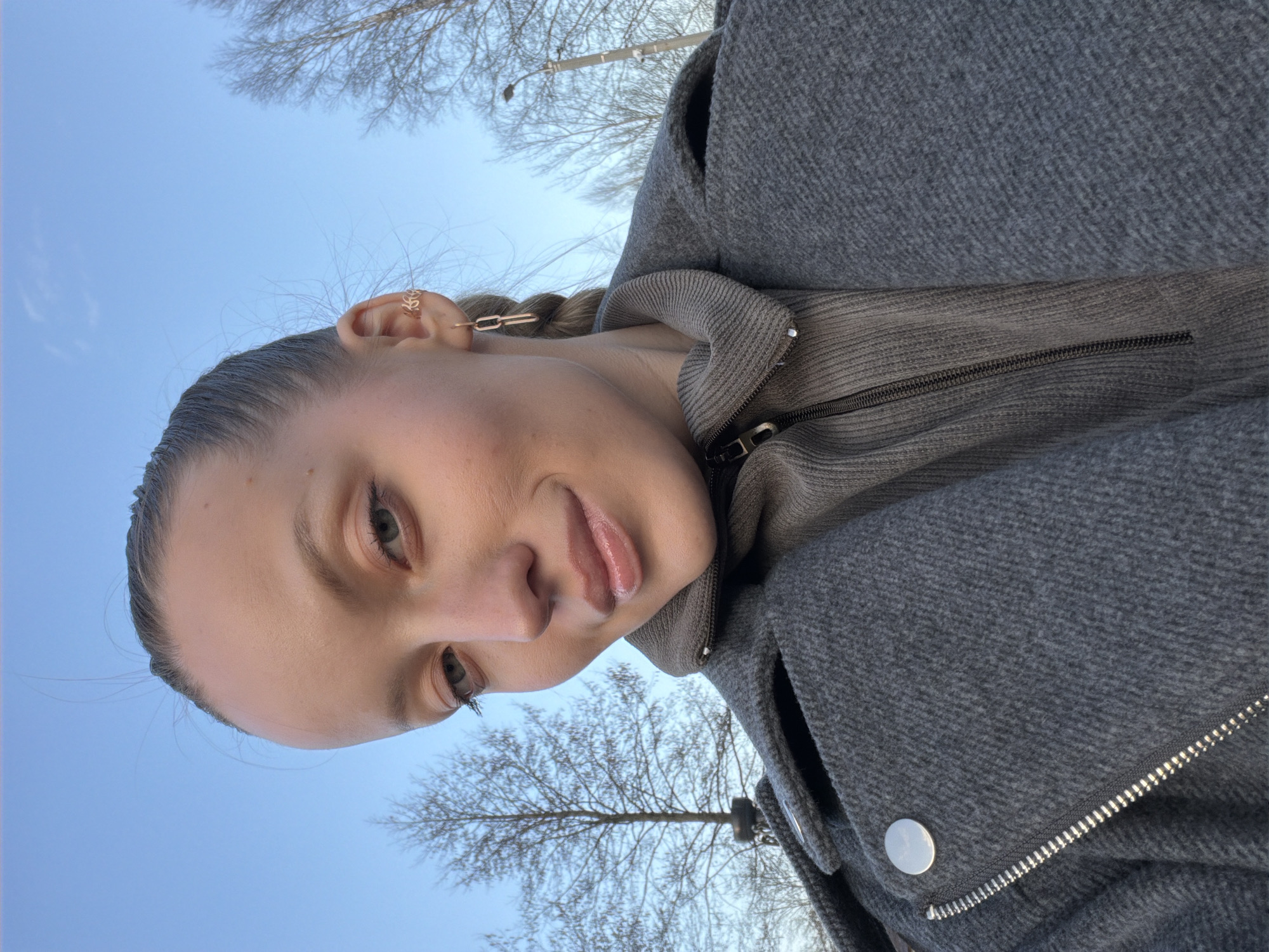 A female subject is captured in a close-up, outdoor selfie photograph in Reshetnikovo, Russia. She has light skin, light-colored eyes, and her hair is pulled back, with a braid visible on the right side. Her expression is a slight smile, with her gaze directed towards the camera. She wears a dark gray, textured, wool-like coat with a wide collar, a silver snap button on the left, and a prominent silver zipper running diagonally across the front. Beneath the coat, a taupe or light brown ribbed knit garment with a high collar and a dark zipper is visible. Her right ear features a gold-colored leaf-motif ear cuff on the upper cartilage and a dangling gold-colored earring composed of two interconnected chain links. The background consists of a clear, bright blue sky and several bare deciduous trees, indicative of a colder season. A dark-colored street lamp is partially visible among the branches in the upper right quadrant of the image. The photo is taken from a slightly low angle, looking upwards.