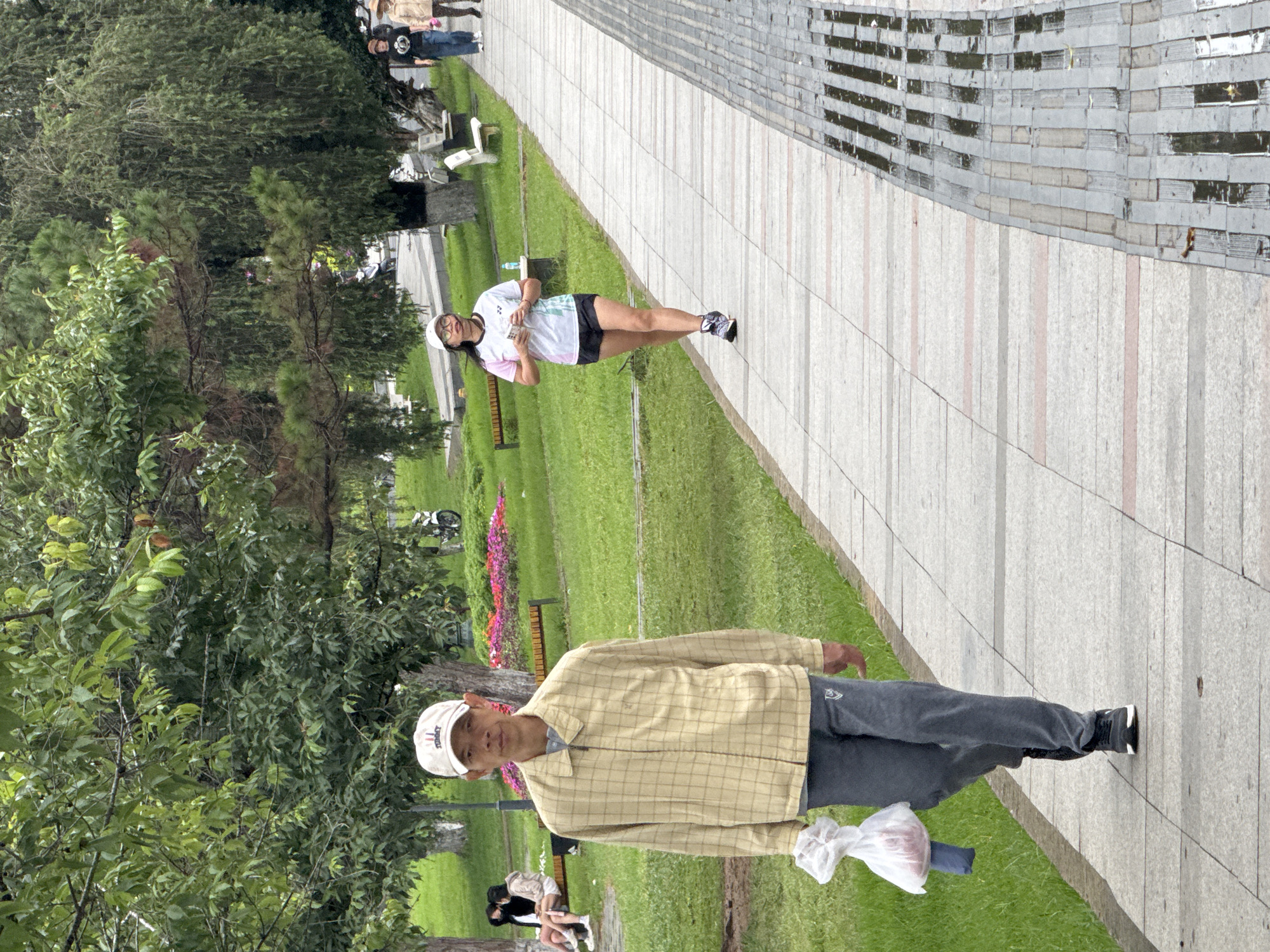 The image captures a vibrant park scene in Da Lat, Vietnam, featuring multiple individuals enjoying the outdoor space. A wide, light-colored paved pathway runs diagonally through the frame, bordered by lush green grass and numerous trees.
In the foreground, an older man is walking towards the viewer on the paved path. He wears a light yellow and brown plaid jacket over a lighter shirt, dark grey pants, and white athletic shoes. A white baseball cap, bearing the partial text "TOMMY," covers his head. He carries a white plastic bag, containing a reddish object, in his left hand.
Further down the path, in the mid-ground, a woman is walking away from the viewer. She is dressed in a white short-sleeved top with pink accents, dark shorts, and purple and white athletic shoes. A white baseball cap similar to the man's is on her head. She holds a small white object, possibly a phone, in her hands.
The background reveals more park activity, with several other people visible, some walking and others seated on benches. Colorful flowering plants add splashes of pink and purple to the green landscape. On the right side of the pathway, a distinctive stone wall or building facade with horizontal grey and lighter-colored stripes is prominent. The overall lighting suggests it is daytime, possibly overcast or late afternoon, with soft shadows. The ground appears slightly damp in areas, hinting at recent rain. The atmosphere is calm and leisurely.