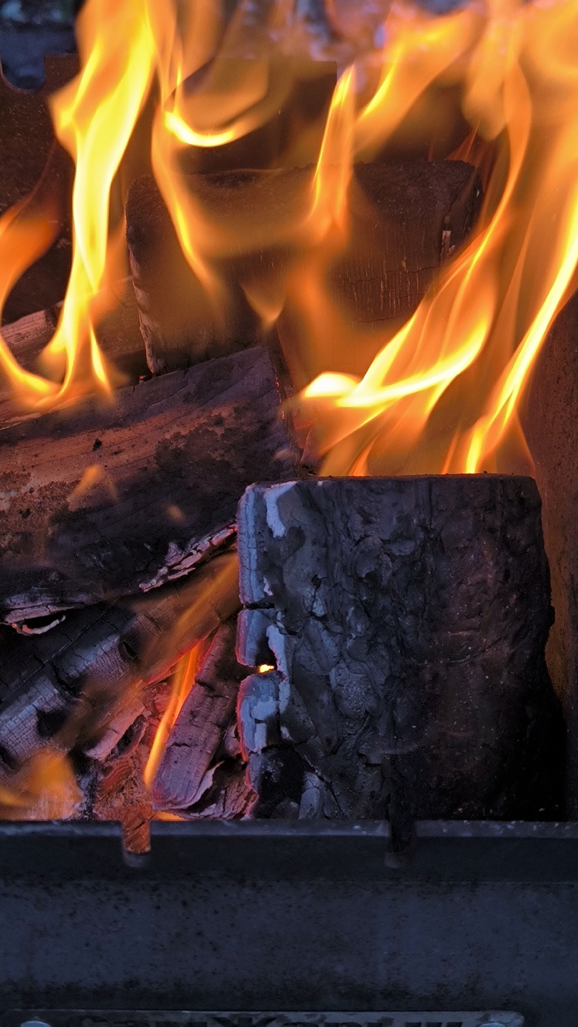 A close-up, vertical, eye-level shot captures a fire burning in a metal brazier. The flames are predominantly bright orange and yellow, with some lighter white accents, flickering and swirling around several pieces of dark, charred wood. One prominent log in the foreground, oriented vertically, has a rough, textured surface and appears partially consumed by the fire, with glowing embers visible within its cracks. Behind it, other pieces of wood, also dark and blackened, are stacked and burning. The background is a dark, indistinct blur. The bottom of the frame shows the upper edge of the dark metal brazier. There are no people present in the image. The scene depicts a typical outdoor fire pit, common in parks or backyards. The location in Miass, Russia, is not visually discernible from the image itself.