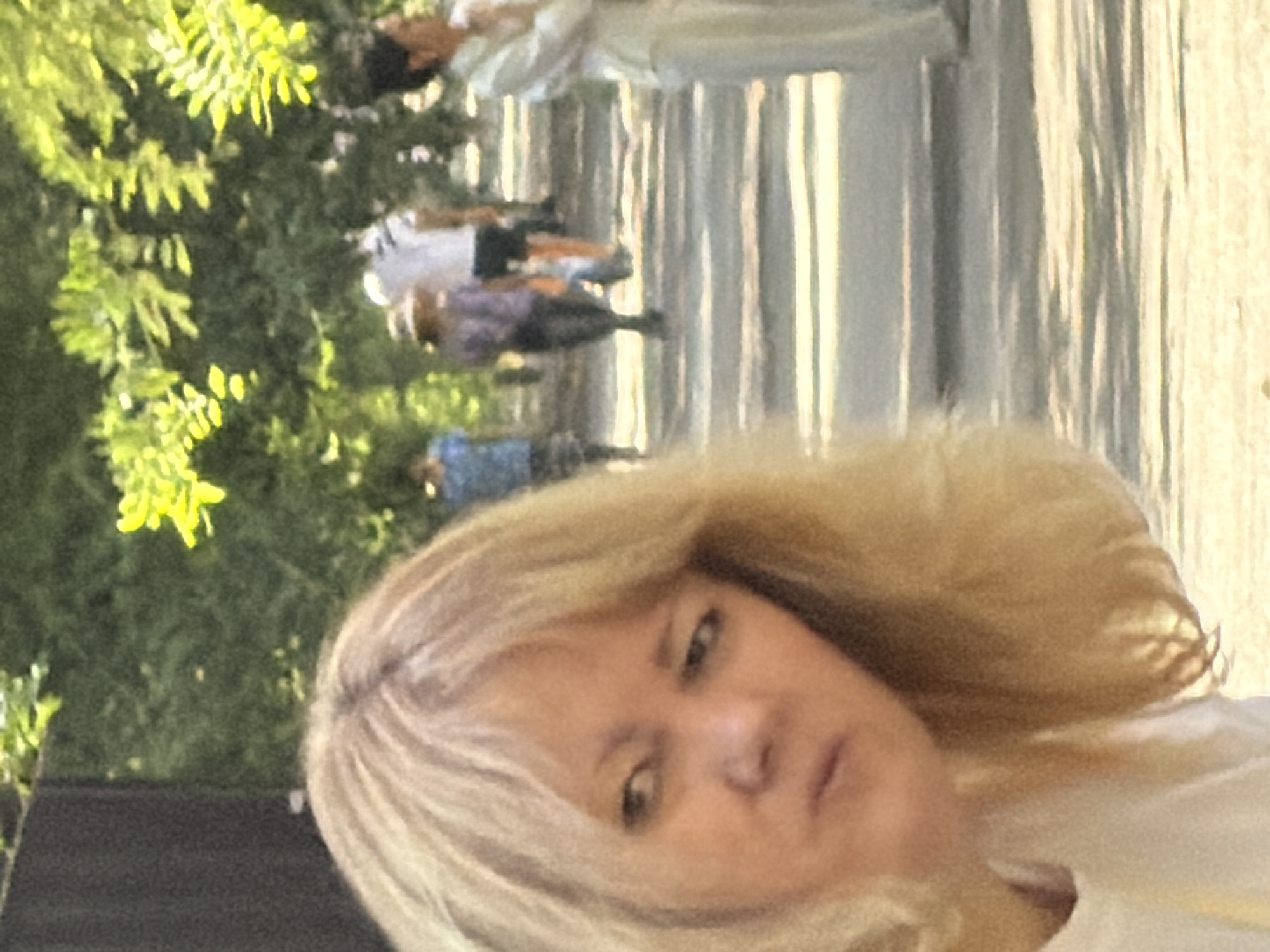 In the foreground of the image, a blonde woman's face is partially visible, looking off to the left with a furrowed brow. Her hair is a light blonde color. Behind her, in the mid-ground, several individuals are walking on a paved path lined with trees. One person in the distance is wearing a white shirt and dark shorts, another is wearing a purple top and dark pants, and a third is wearing a blue shirt and a patterned skirt. Further in the background, a person in a white outfit is visible. The environment appears to be an outdoor setting with lush green foliage and dappled sunlight on the path. The lighting suggests it is daytime.