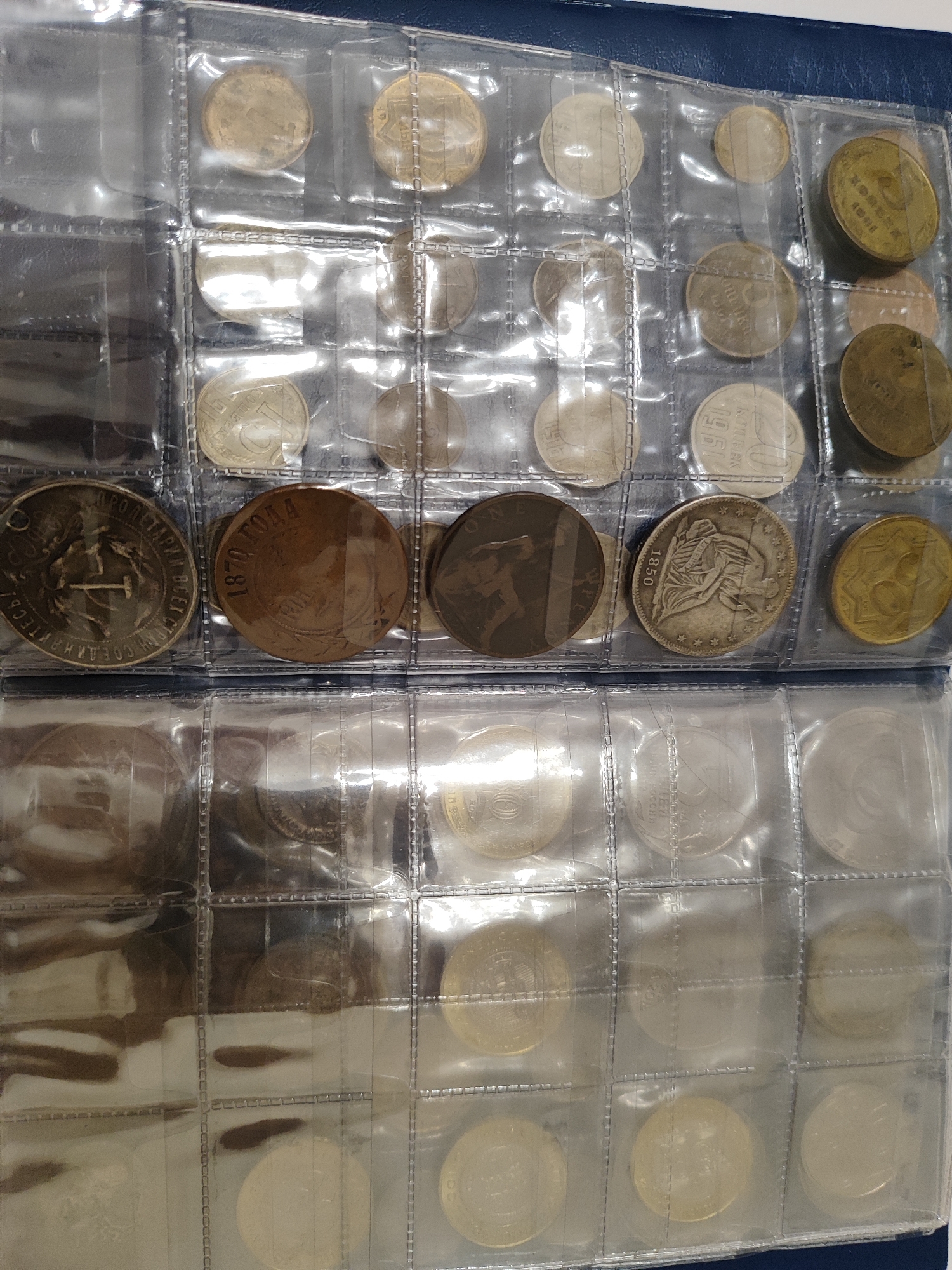 The image is a close-up, overhead view of a coin collector's album. The album is blue and contains multiple clear plastic pages, each divided into rectangular slots holding various coins. The coins are of different sizes, colors, and designs, appearing to be a mix of modern and older currency. Many of the coins have visible text and imagery, such as dates and portraits. The lighting suggests an indoor environment. There are no people visible in the image, and no specific geographic location, such as the city of Pavlodar, Kazakhstan, is depicted or implied by the contents of the album.