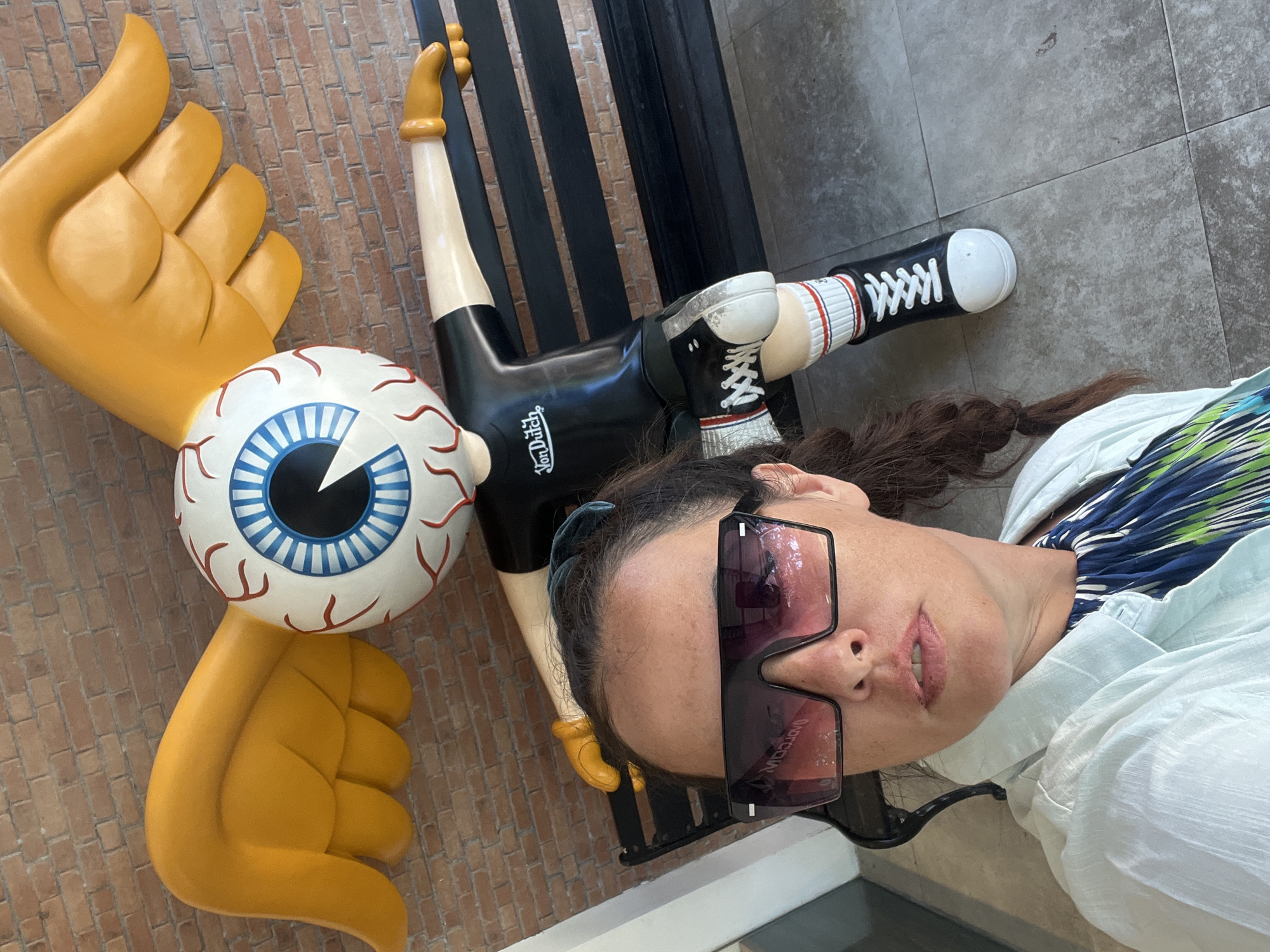 A woman takes a selfie in front of a statue of a character with a large eyeball for a head, yellow wings, and a black and white outfit, located in Kuta Selatan, Indonesia. The statue is seated on a black bench. The woman is wearing large, dark sunglasses, a light blue shirt, and a blue, green, and white patterned dress. The background consists of a brick wall and tiled flooring.