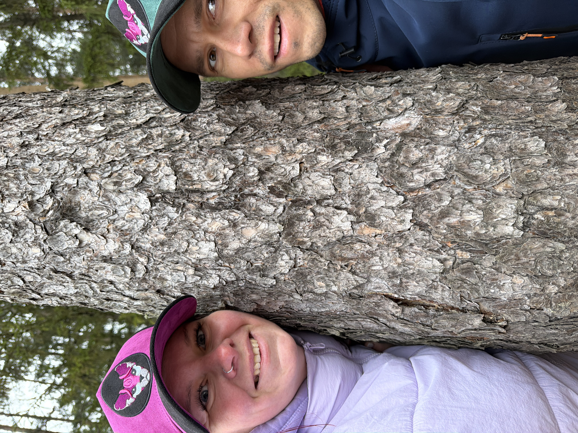 Two people, a man and a woman, are lying on either side of a large tree trunk. The woman is wearing a pink cap and a light purple jacket, while the man is wearing a green and black cap and a blue jacket. Both are smiling. The location appears to be a forest or wooded area. The image was likely taken in Chaikovsky, Russia.