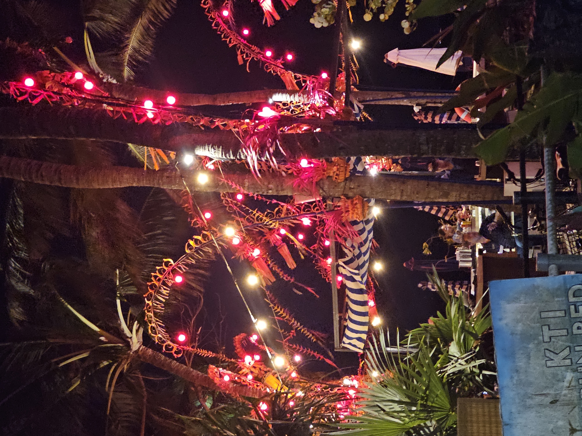 The image depicts a nighttime scene at a tropical venue, likely a beachside bar or restaurant, in Denpasar, Indonesia. The foreground is dominated by lush green palm fronds and a weathered blue sign with partial white lettering that reads "KTI". Beyond this, the scene opens up to an area adorned with numerous red and yellow string lights that illuminate palm trees and decorative elements. A striped awning, in blue and white, is visible above a wooden bar area. Several people are present, appearing to be patrons and staff. One individual in the middle ground is seated at the bar, while others are standing or interacting in the background. An umbrella stands near the right side of the frame. The overall atmosphere suggests a lively outdoor gathering, possibly in a tourist-oriented location.