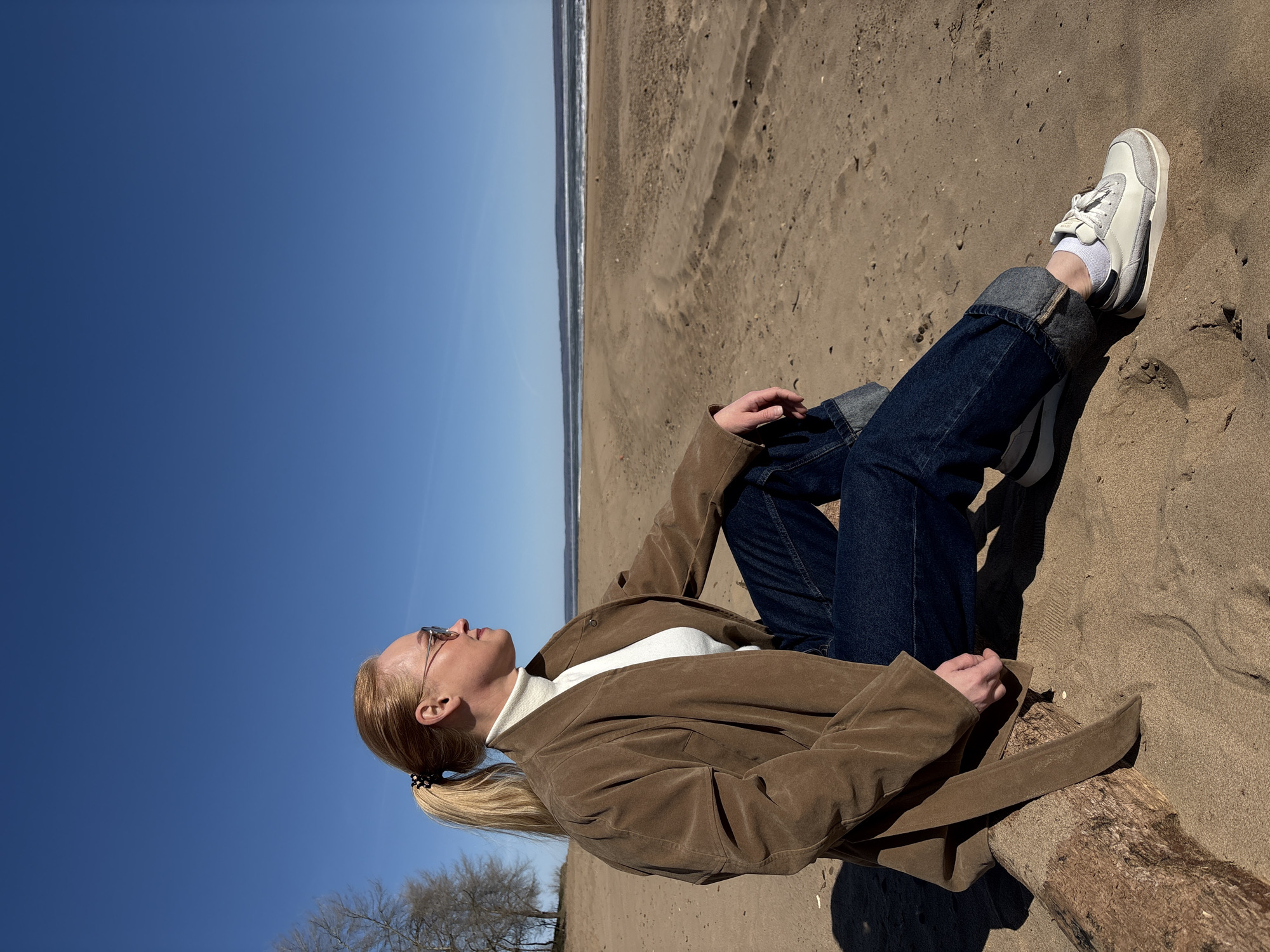 This image captures a blonde individual sitting on a sandy beach under a clear, vast blue sky, characteristic of Noviy, Russia. The composition is uniquely tilted, presenting the horizon line vertically along the left side, dividing the deep blue sky from the sandy shore and ocean.
The person, viewed in profile, is looking upwards, suggesting a moment of relaxation or contemplation. Their blonde hair is pulled back into a ponytail with a patterned scrunchie, and they wear sunglasses. Dressed in a white turtleneck, a brown long-sleeved jacket (possibly suede or corduroy), dark blue cuffed jeans, and white sneakers with white socks, they appear comfortably attired for the outdoors. Their left hand rests casually on their knee.
The setting is a wide, sandy beach. A piece of driftwood or a log is visible near the person's feet and beneath their jacket, casting a distinct shadow. In the distance, the calm ocean can be seen with gentle waves breaking on the shore. The sky is strikingly clear and cloudless, indicating a bright and sunny day. Some bare tree branches are visible in the lower left corner, suggesting either late winter or early spring. The strong sunlight creates sharp shadows across the sand and on the person's clothing. Although the sneakers appear to have a brand logo or text on their side, it is too indistinct to be read.