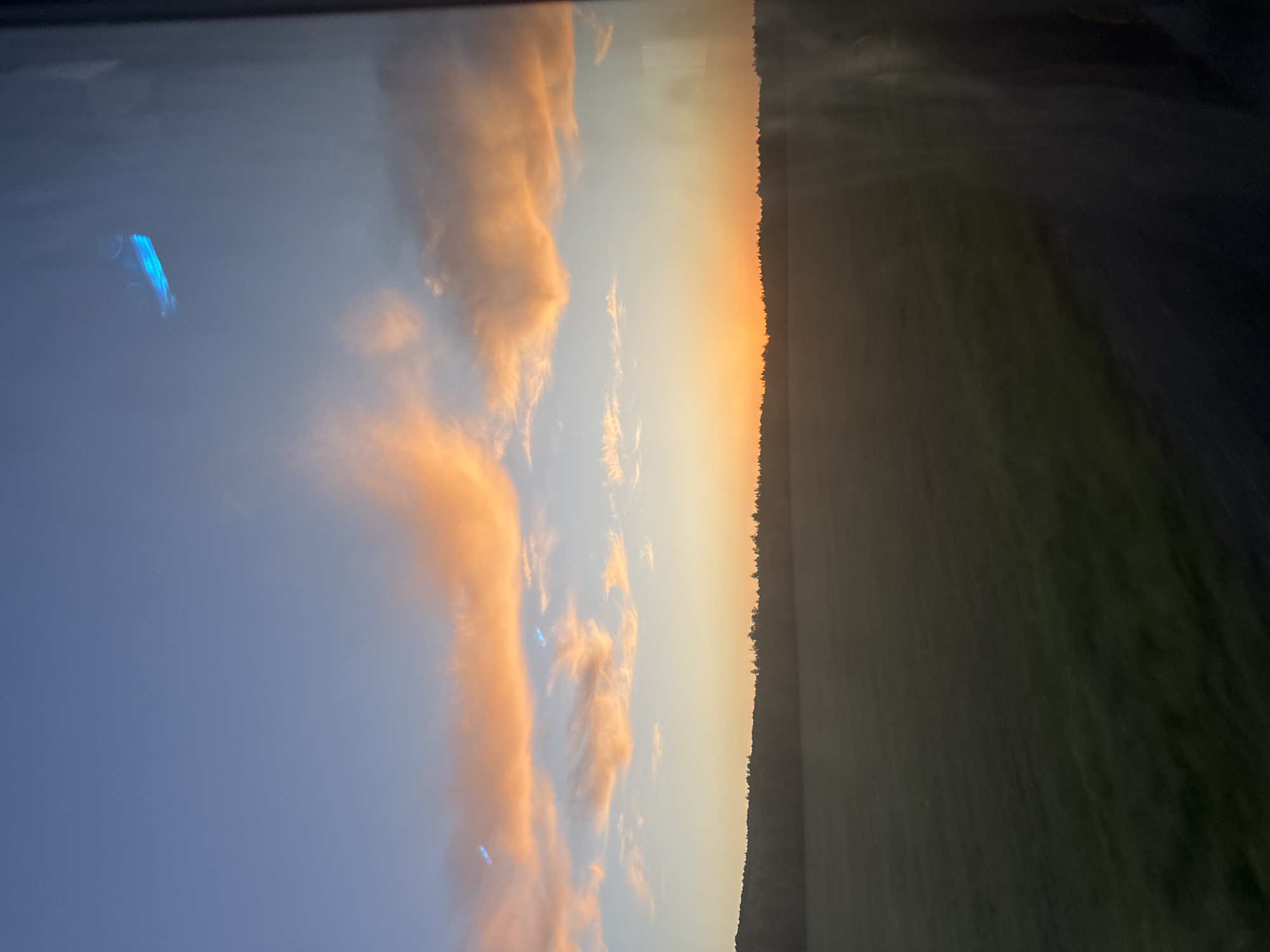 The image shows a landscape at sunset, possibly taken through a window. On the left, there is a sky with clouds tinted orange by the setting sun. A reflection, possibly of a car dashboard, is visible in the upper left corner. The horizon line is marked by trees. To the right of the trees, a dark green field or area of vegetation is present. The overall impression is that the photograph was taken in a rural setting, possibly from inside a vehicle or building. The location, Yasashnaya Tashla, Russia, cannot be independently verified from the image alone.